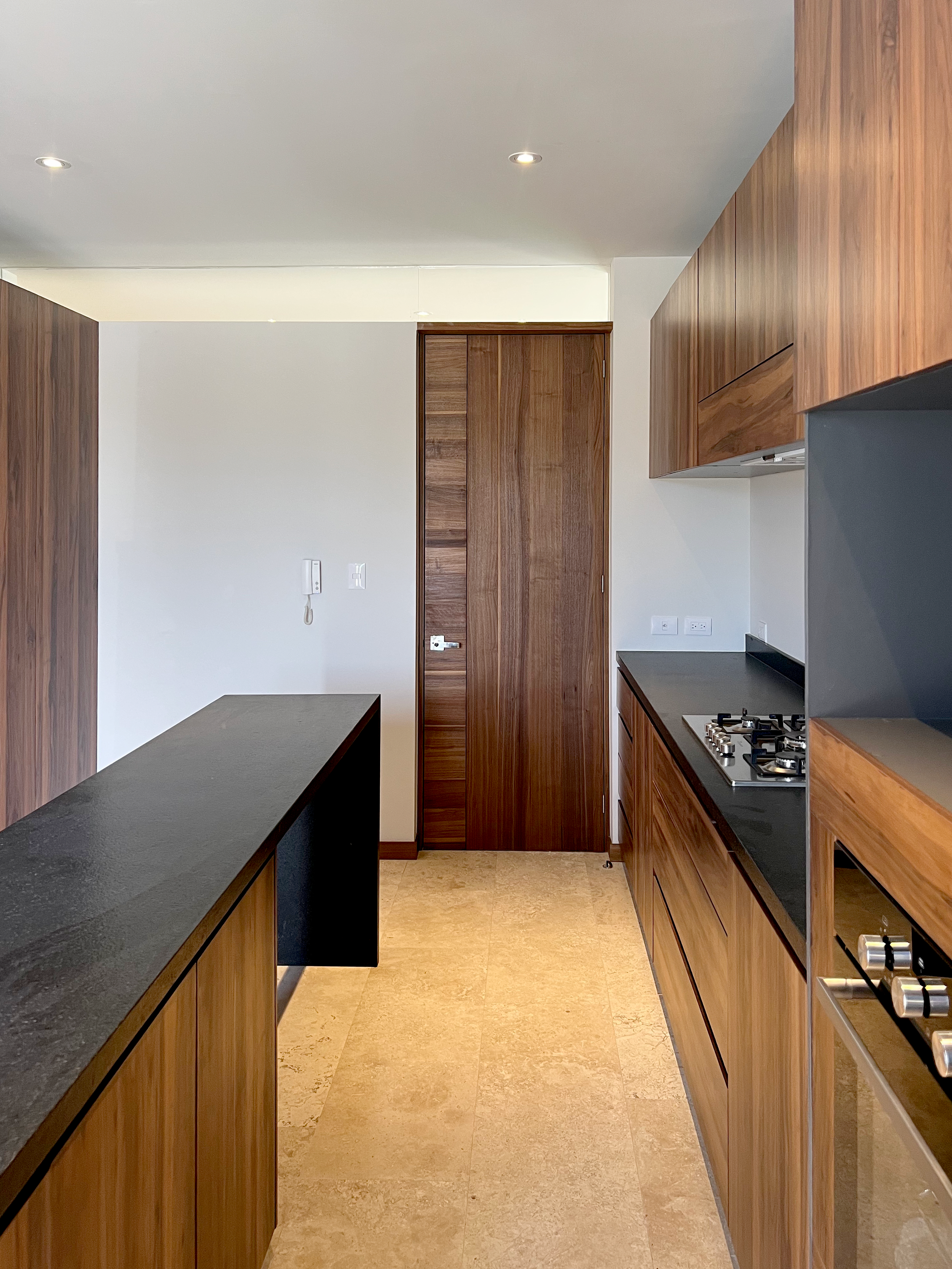 Cocina moderna con gabinetes de madera, encimera negra, estufa de gas, paredes blancas y suelo de piedra beige.