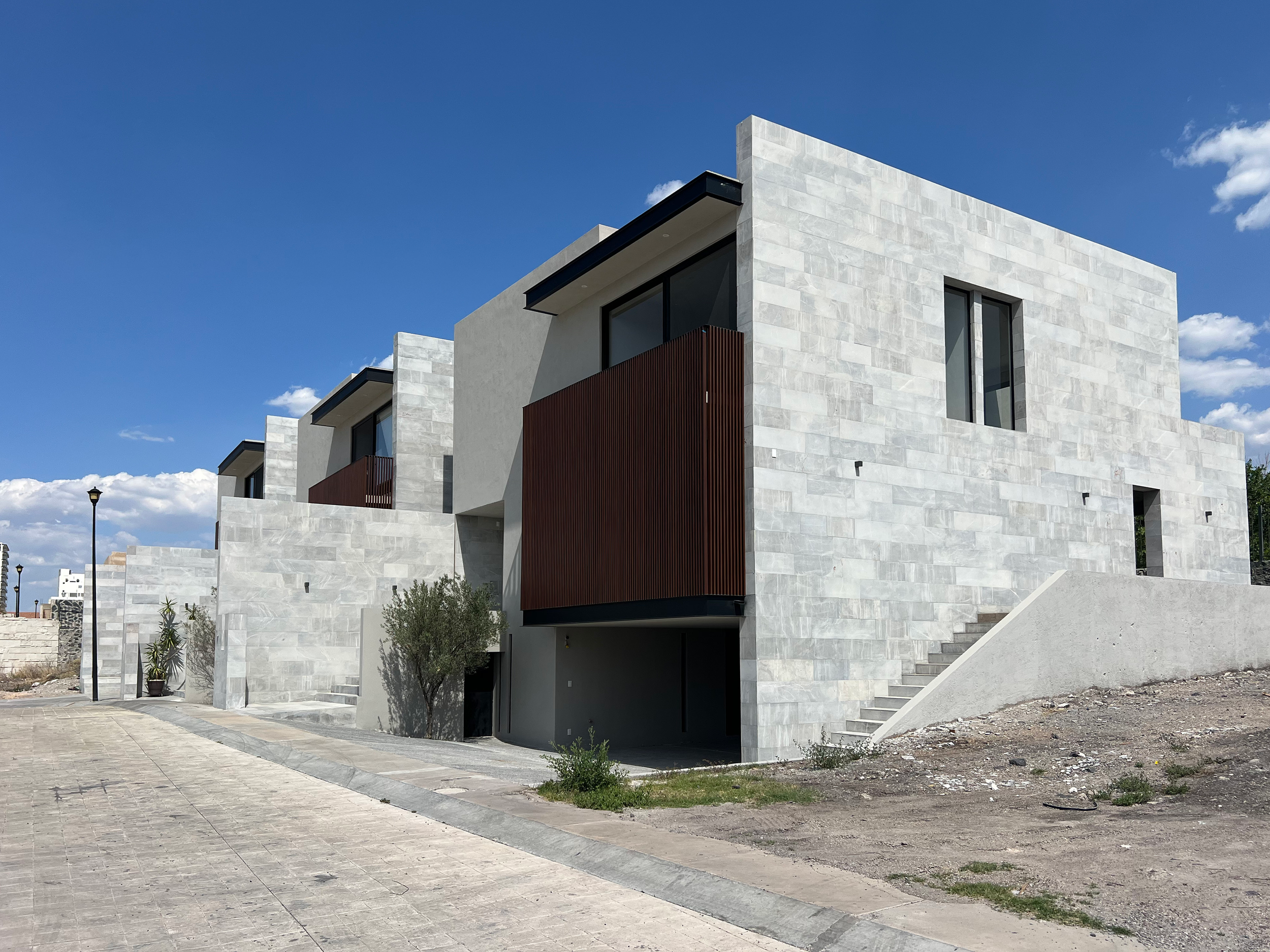 Edificio moderno de varias plantas con fachada de piedra clara, ventanas grandes, y una estructura de madera en una sección. Está en un entorno con camino de piedra y se observa un cielo azul con algunas nubes.