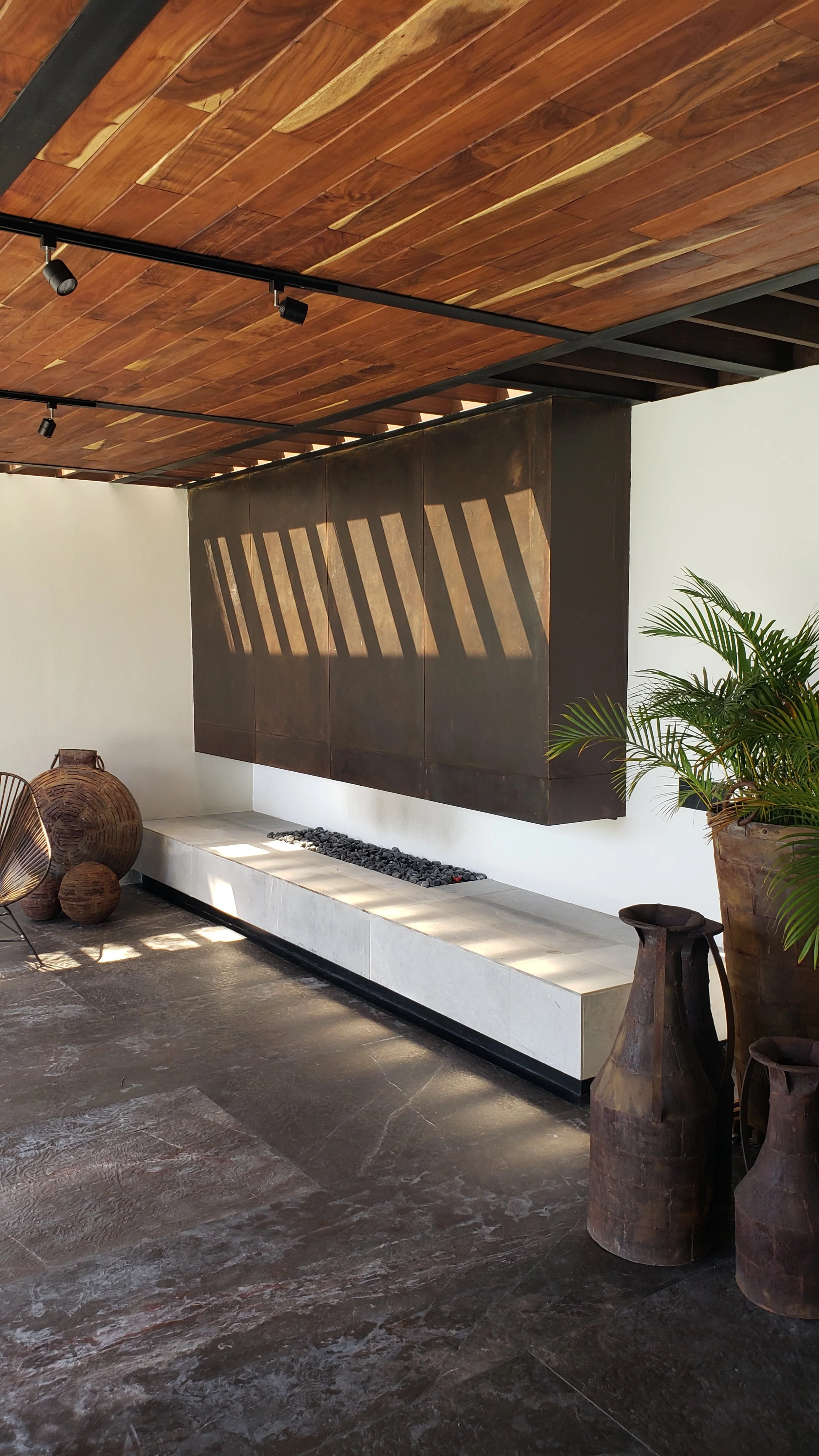 Espacio interior con techo de madera, paredes blancas, una chimenea de metal oscura, plantas en macetas grandes y decorativas, y terreno de piedra uniforme.