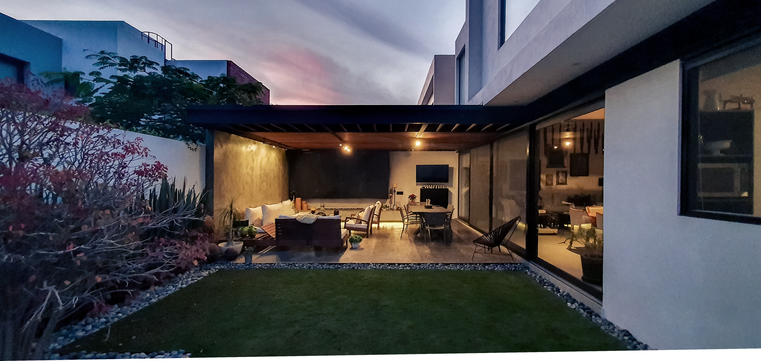 Vista de un patio moderno al atardecer, con áreas de descanso y una pantalla de televisión en la pared trasera, rodeado de plantas y ventanas grandes que reflejan la luz interior.