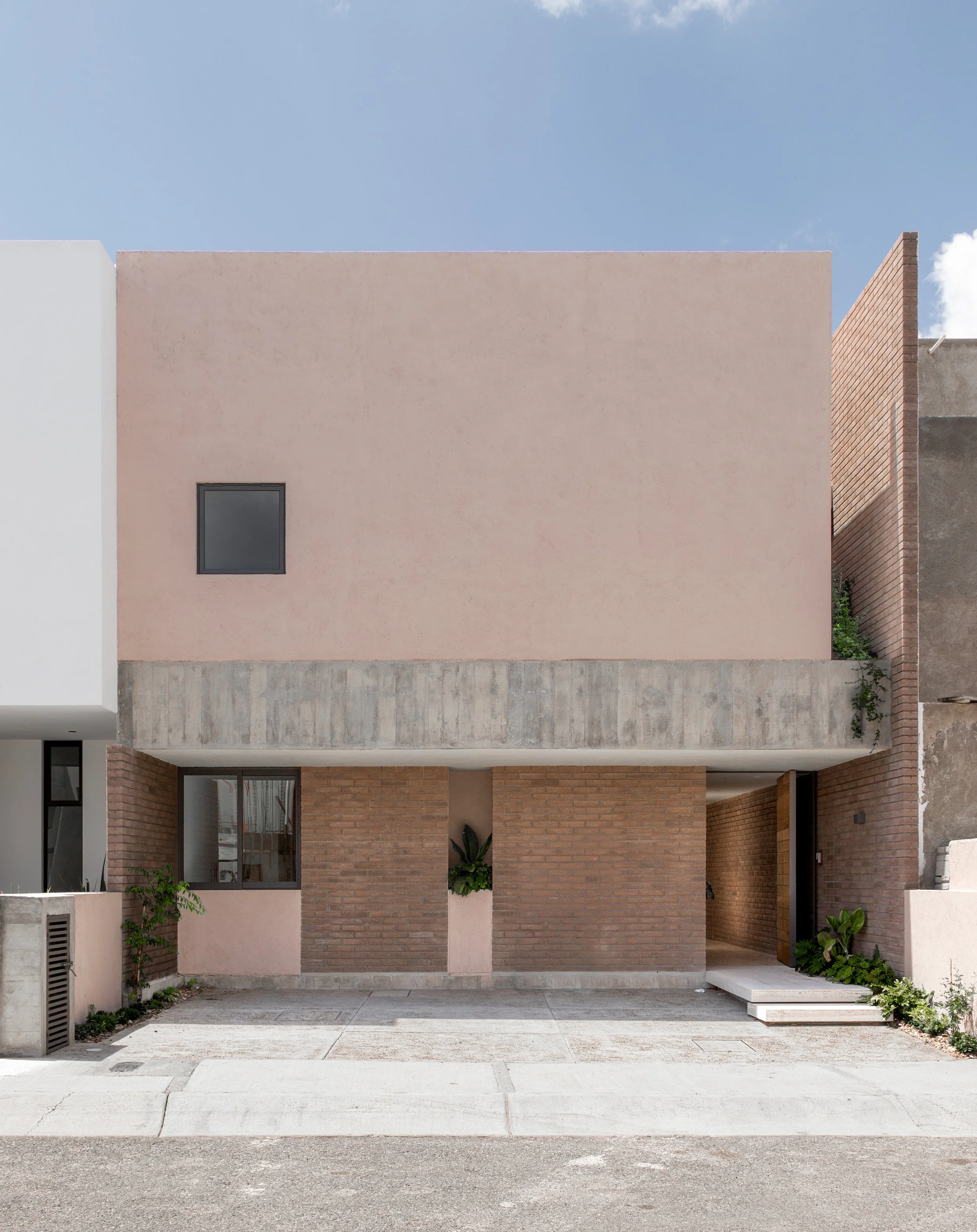 Edificio moderno con fachada de ladrillos, ventanas pequeñas y plantas decorativas, en un día soleado con cielo azul.