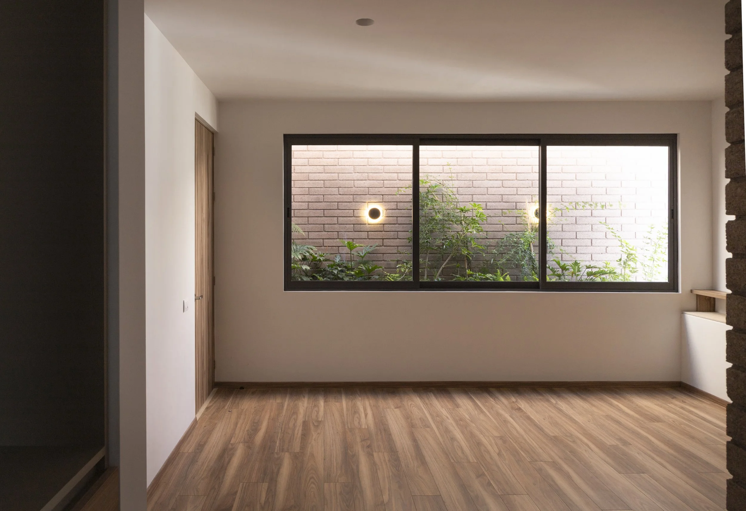 Sala vacía con ventana grande, vista a jardín con plantas y muro de ladrillos, suelo de madera y pared blanca.
