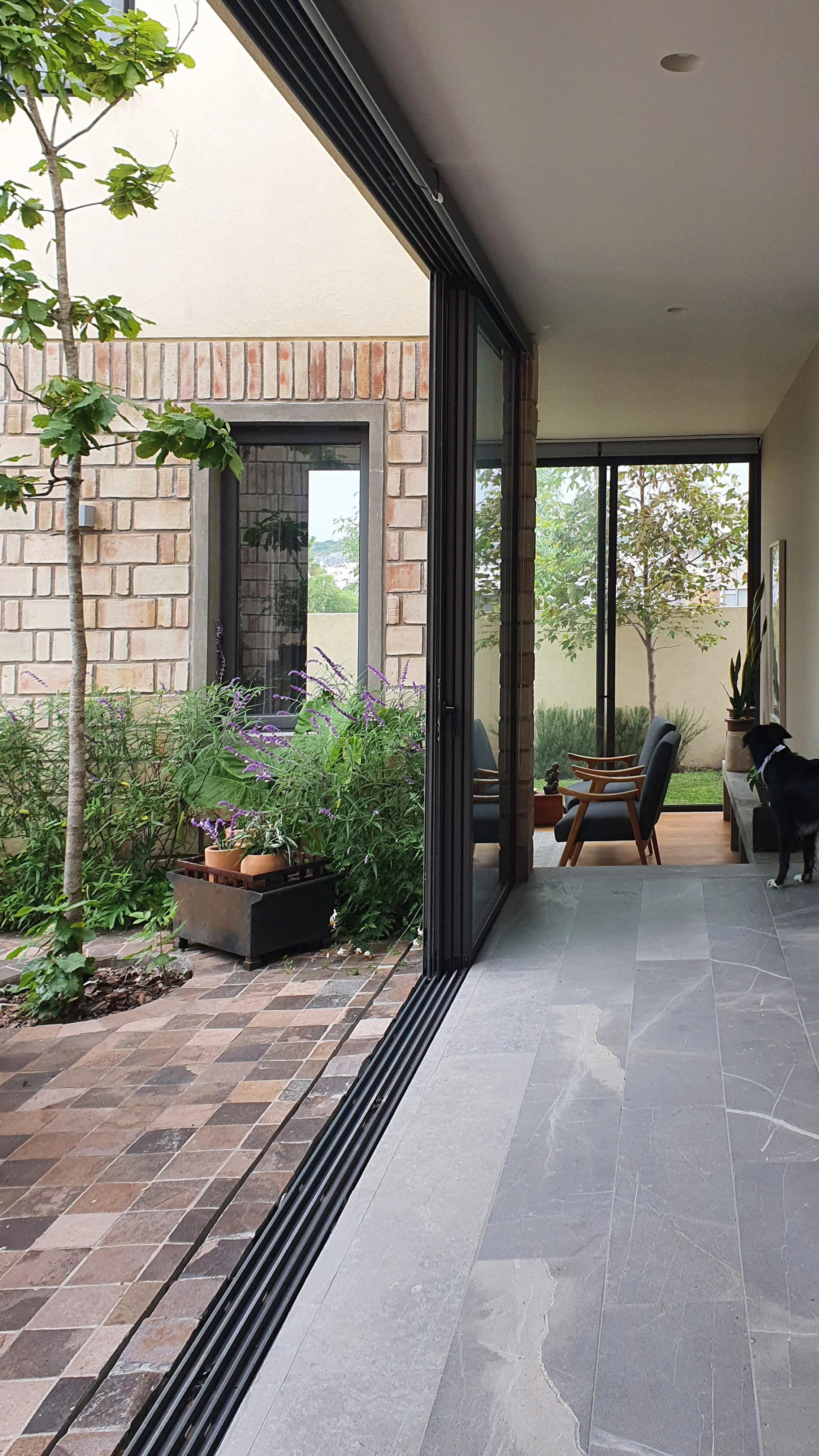 Visión interior de un hogar con puertas corredizas de vidrio que reflejan un área de patio con plantas, un árbol y una pared de ladrillos. Dentro, hay sillones con madera y cojines junto a una televisión, y en el suelo hay baldosas de piedra gris.