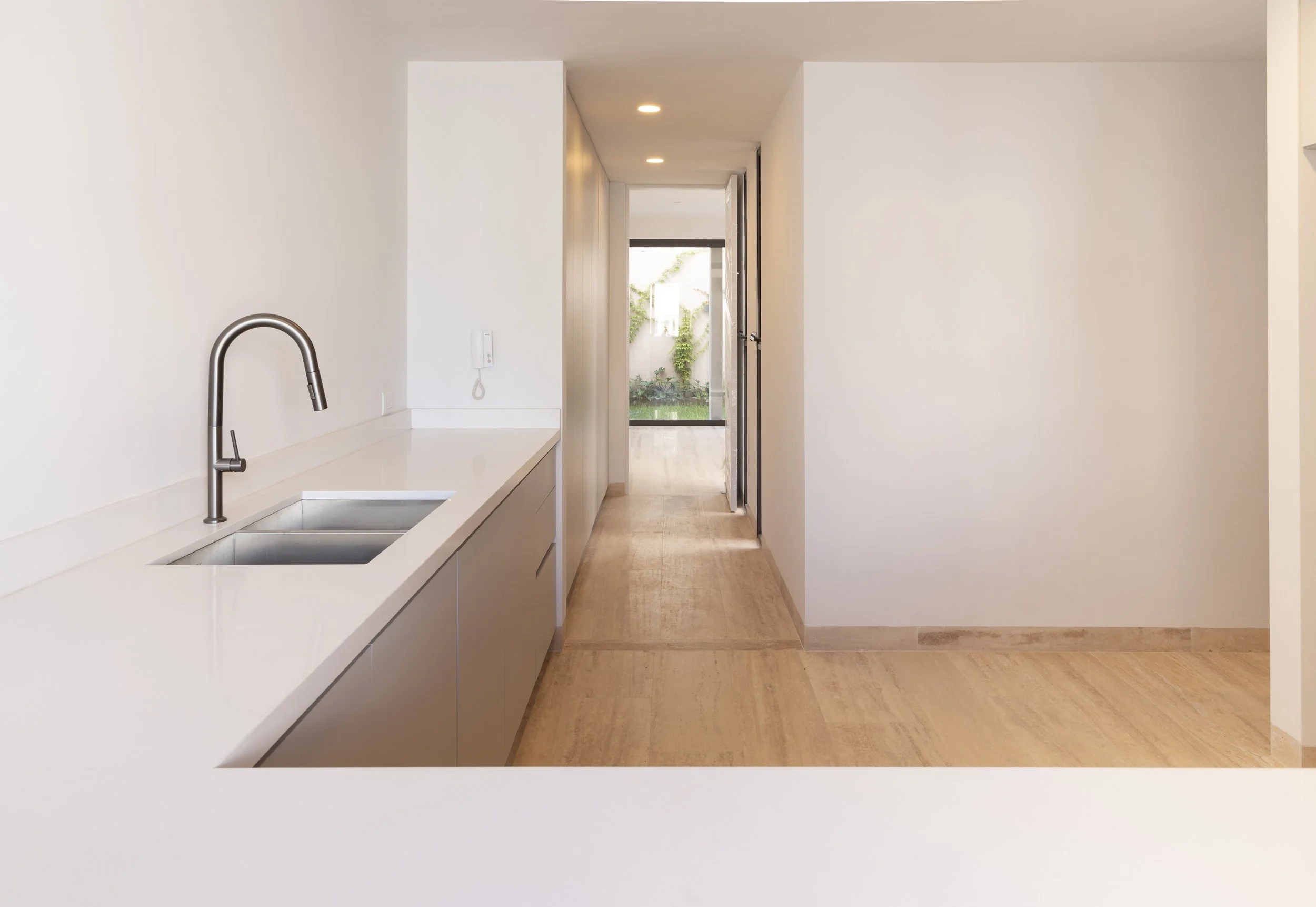 Cocina moderna con encimera blanca, lavabo y grifo de acero inoxidable, pasillo que lleva a un área con puerta de cristal y jardín al fondo.