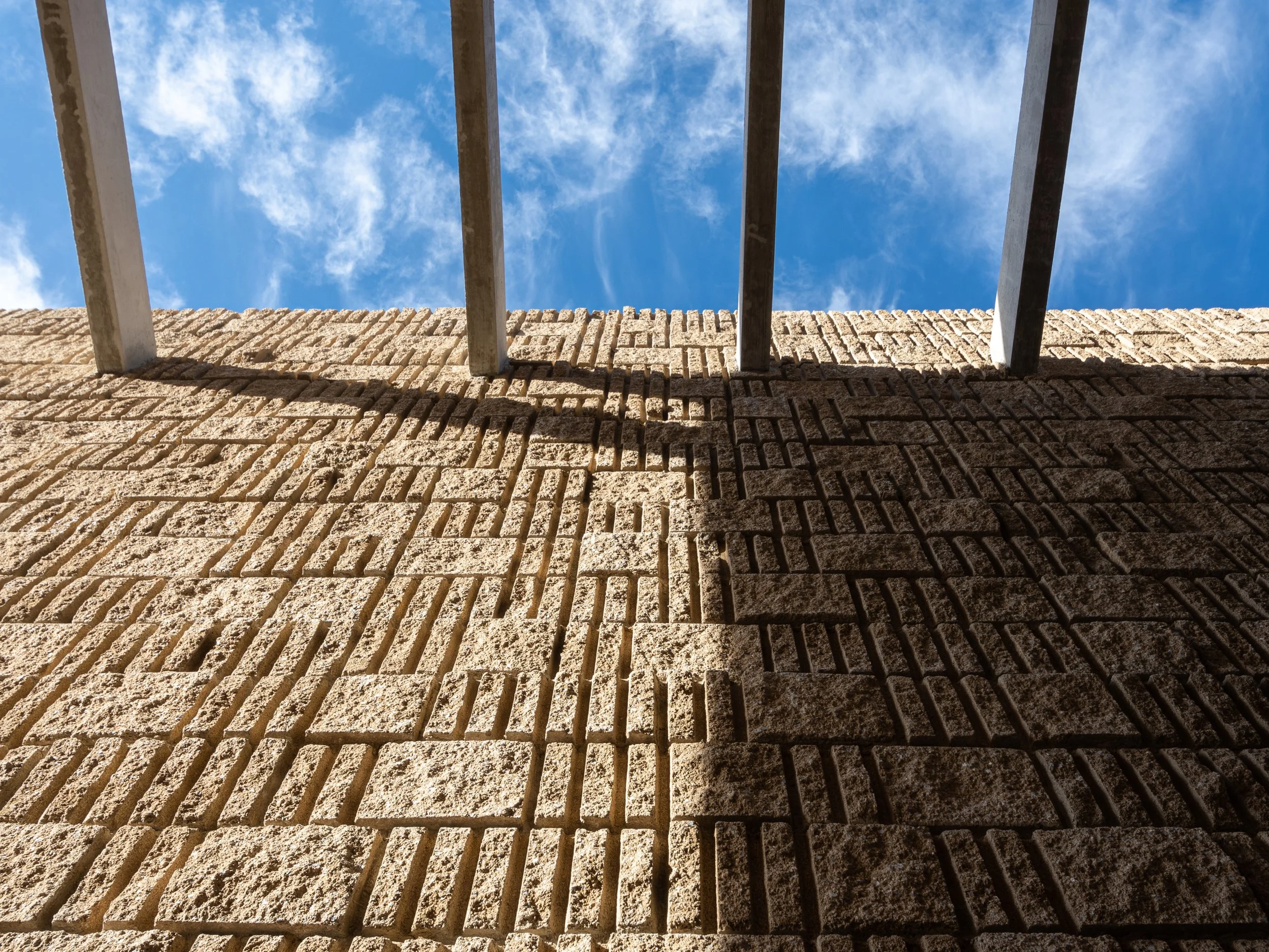 Vista desde abajo de un muro de ladrillos con una reja metálica en la parte superior, cielo con nubes al fondo.
