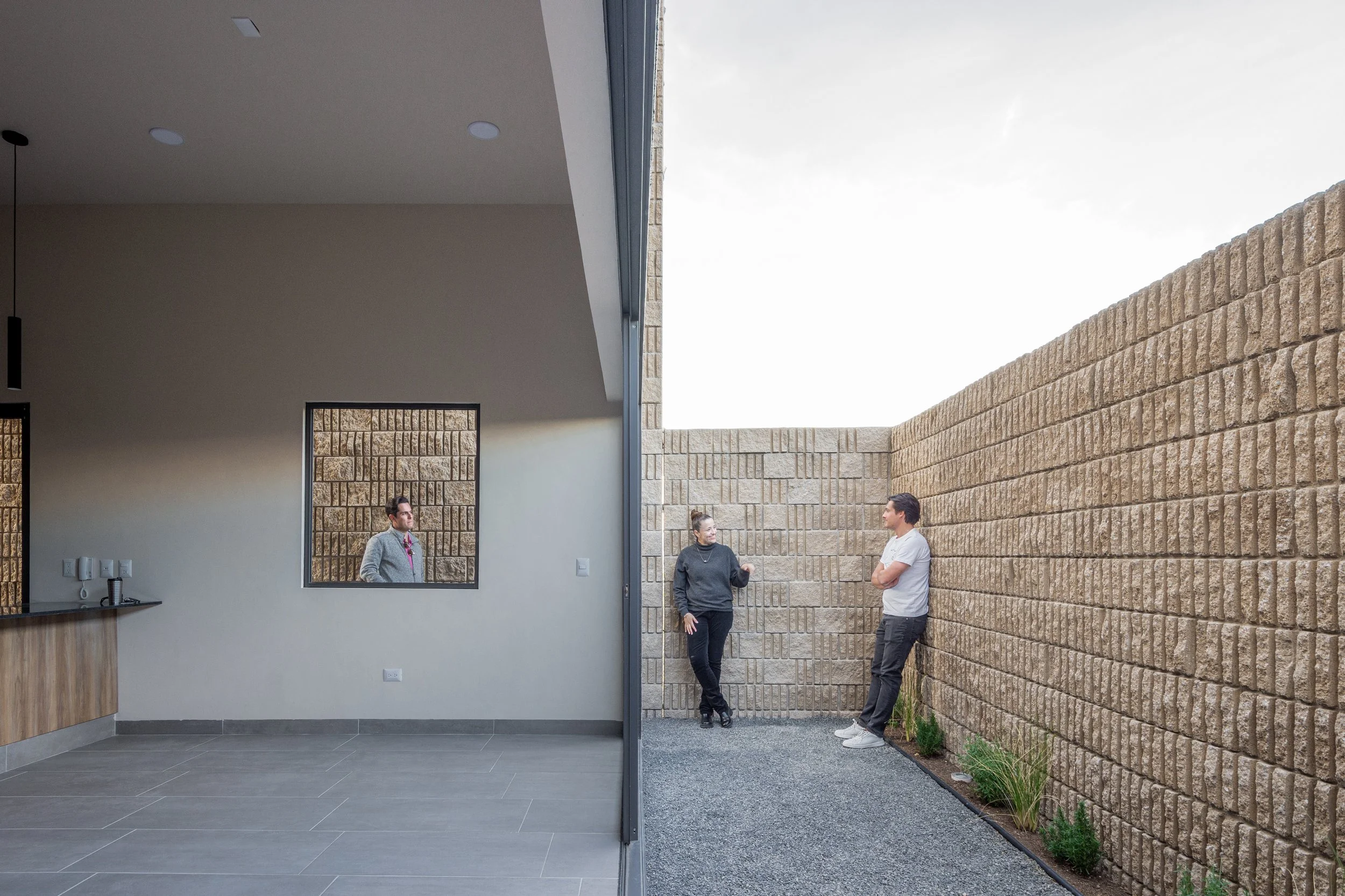 Escena que muestra una transición de un espacio interior a uno exterior, con dos personas conversando en el patio, y una tercera persona visible a través de una ventana interior.