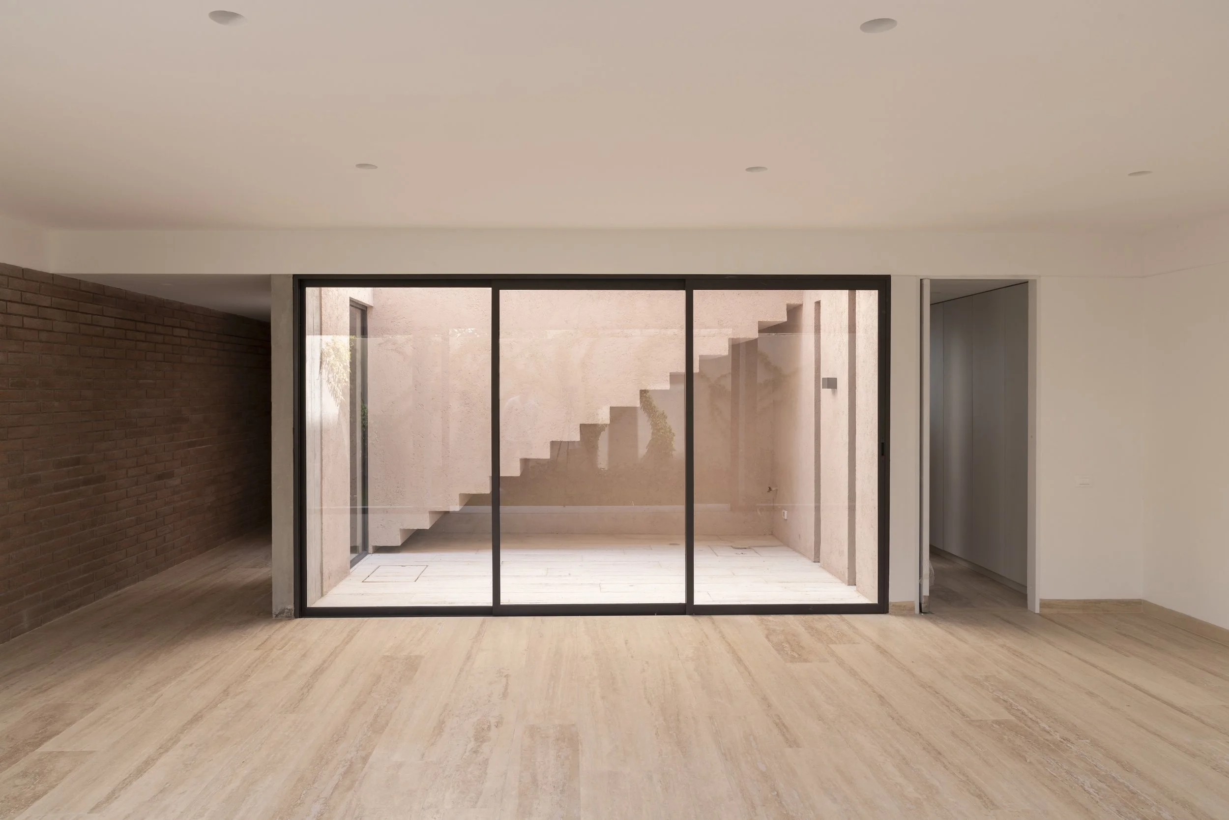 Espacio interior con suelo de madera, pared de ladrillos en un lado, y una pared blanca. Ventana grande de vidrio que da a un patio con escaleras de concreto.