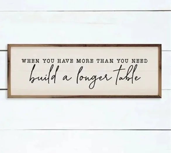 A framed wall sign with the quote, 'When you have more than you need, build a longer table,' hanging on a white shiplap wall.