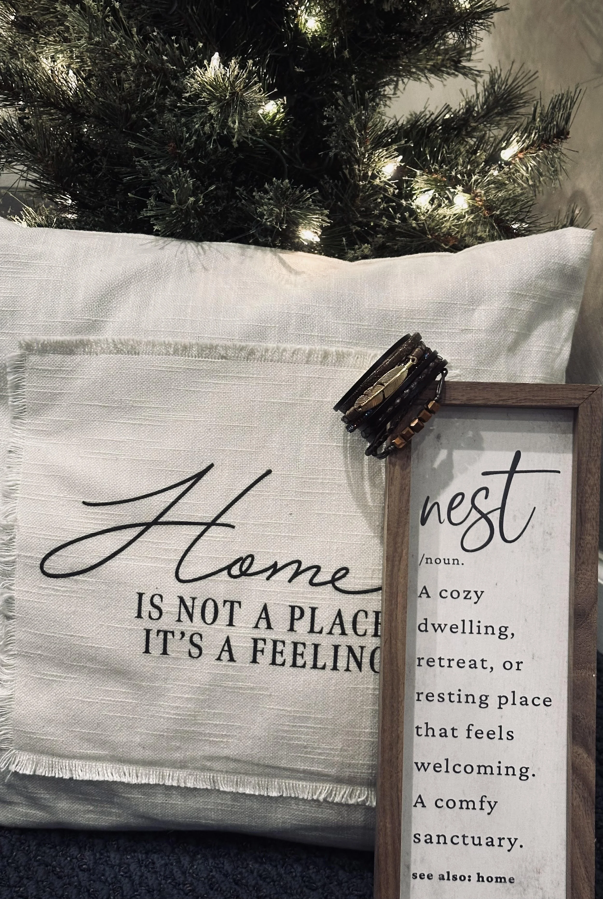 Decorative image featuring a white pillow with black text that says, 'Home is not a place, it's a feeling,' and a framed sign with the word 'nest' and a definition, placed in front of a Christmas tree with lights, along with some bracelets draped over the top of the frame.