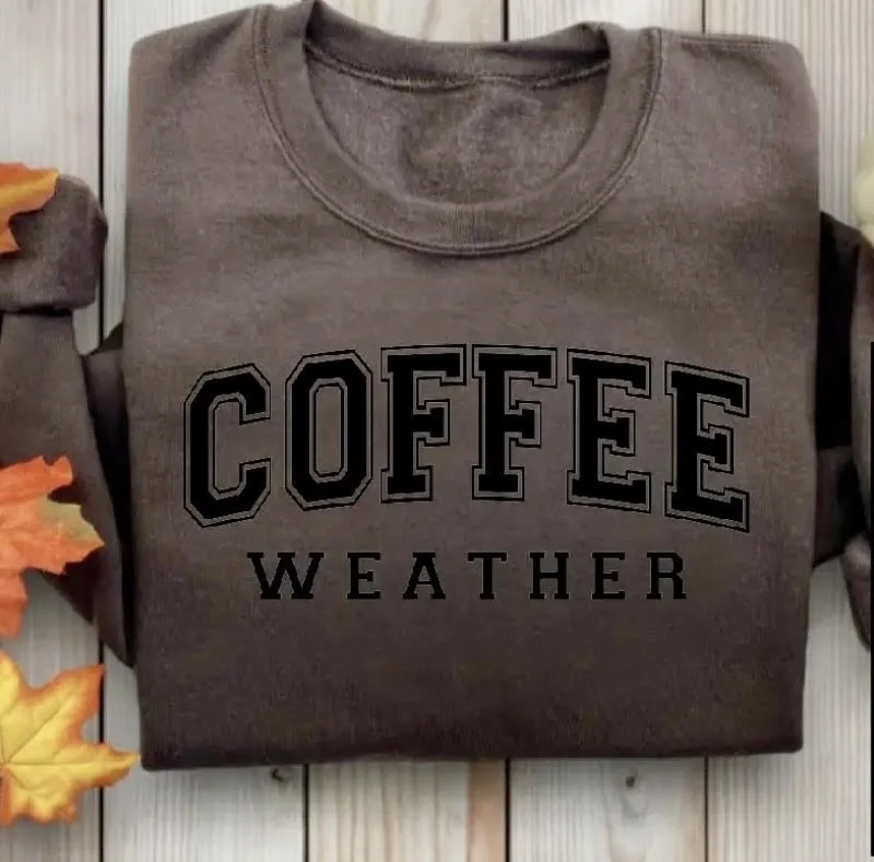 Gray sweatshirt with the words "Coffee Weather" printed on the front, laid out on a wooden surface with autumn leaves nearby.