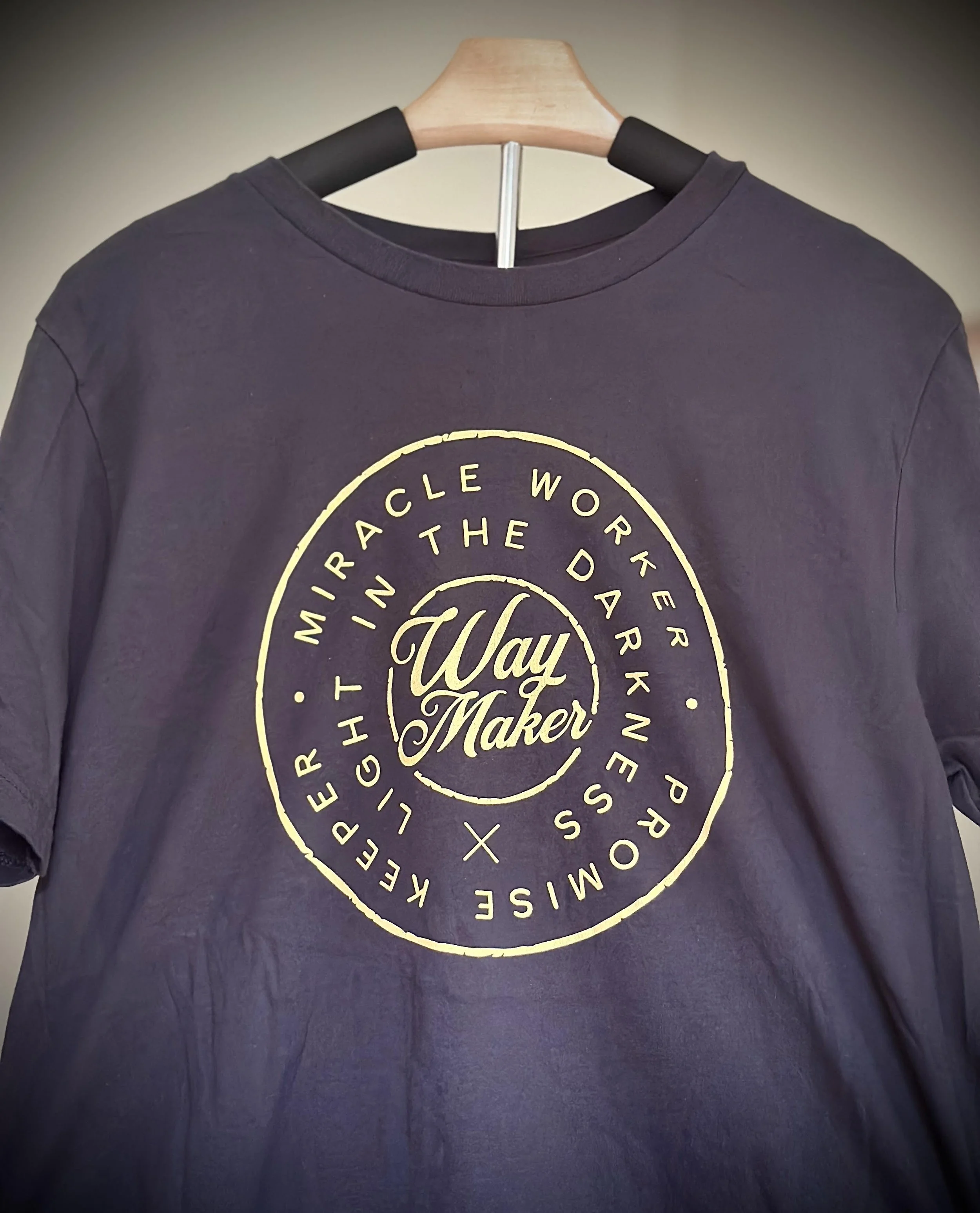 Black T-shirt, with gold circular text that reads 'Way Maker' in the center and 'Miracle Worker, Light in the Dark, Never Promises, Kept the Faith' around the circle.