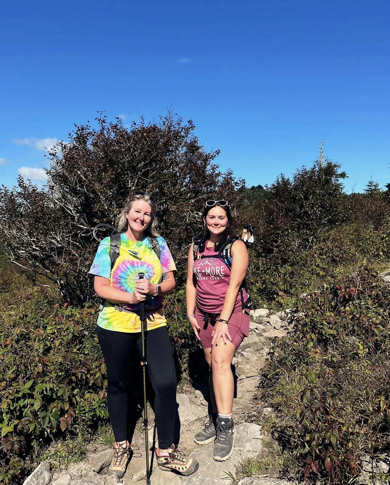 Two women hiking on a rocky trail surrounded by bushes with a clear blue sky overhead.