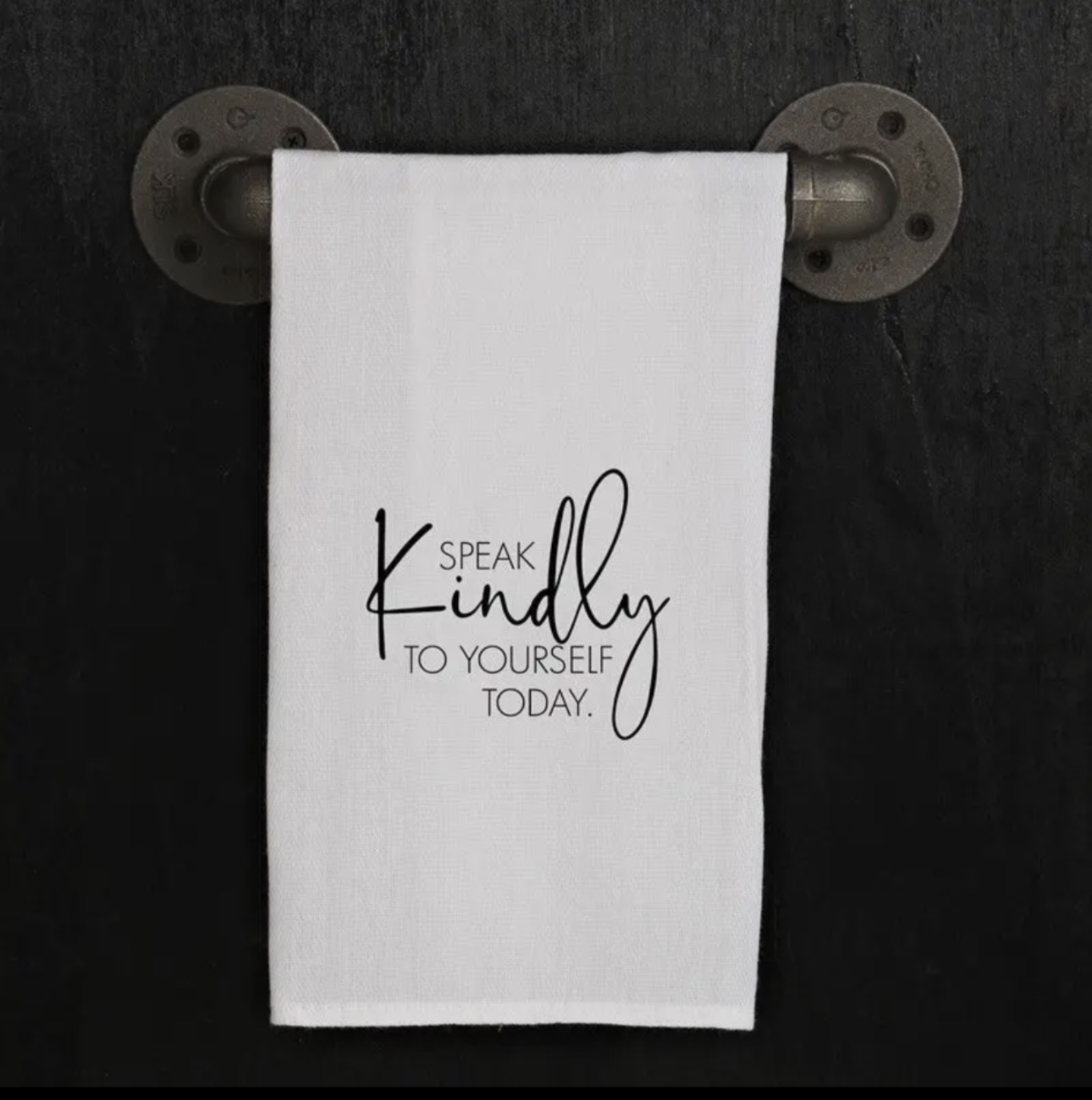 A white towel with the quote 'Speak kindly to yourself today' hanging on a metal rod mounted on a dark wall.