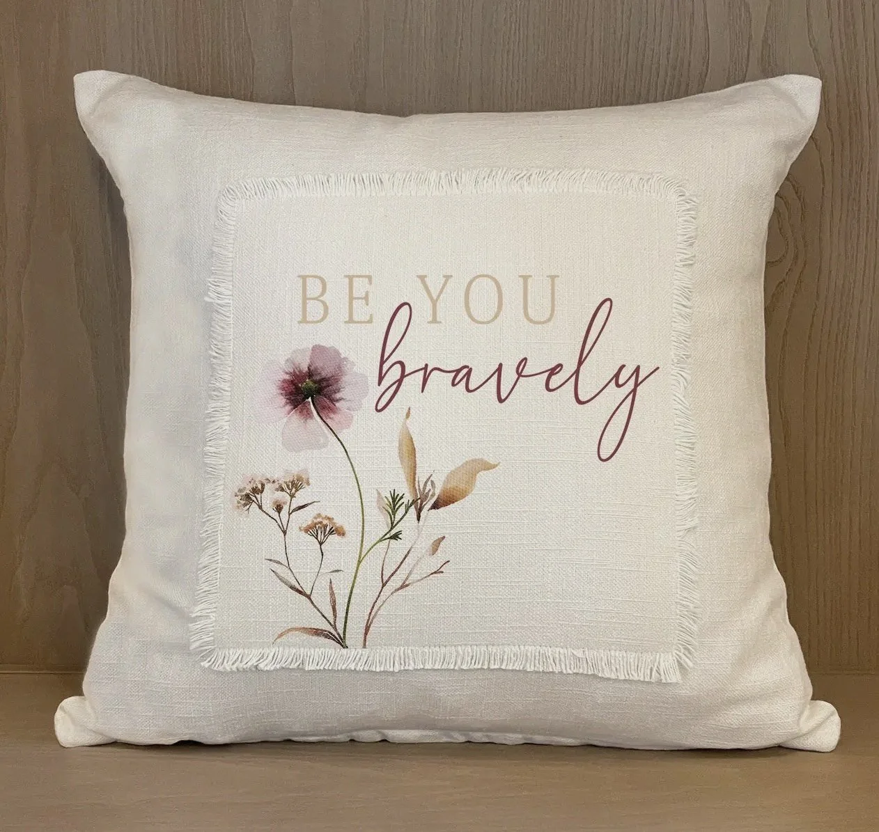 Decorative white throw pillow with a floral design and the phrase 'Be you bravely' printed on it, placed on a wooden surface.