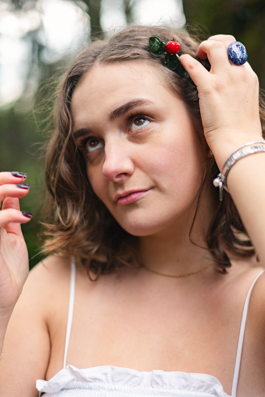 Mulher de cabelo castanho com um enfeite de folhas verdes e uma cereja na cabeça, olhando para cima com expressão pensativa, com mãos próximas à cabeça, ao ar livre.