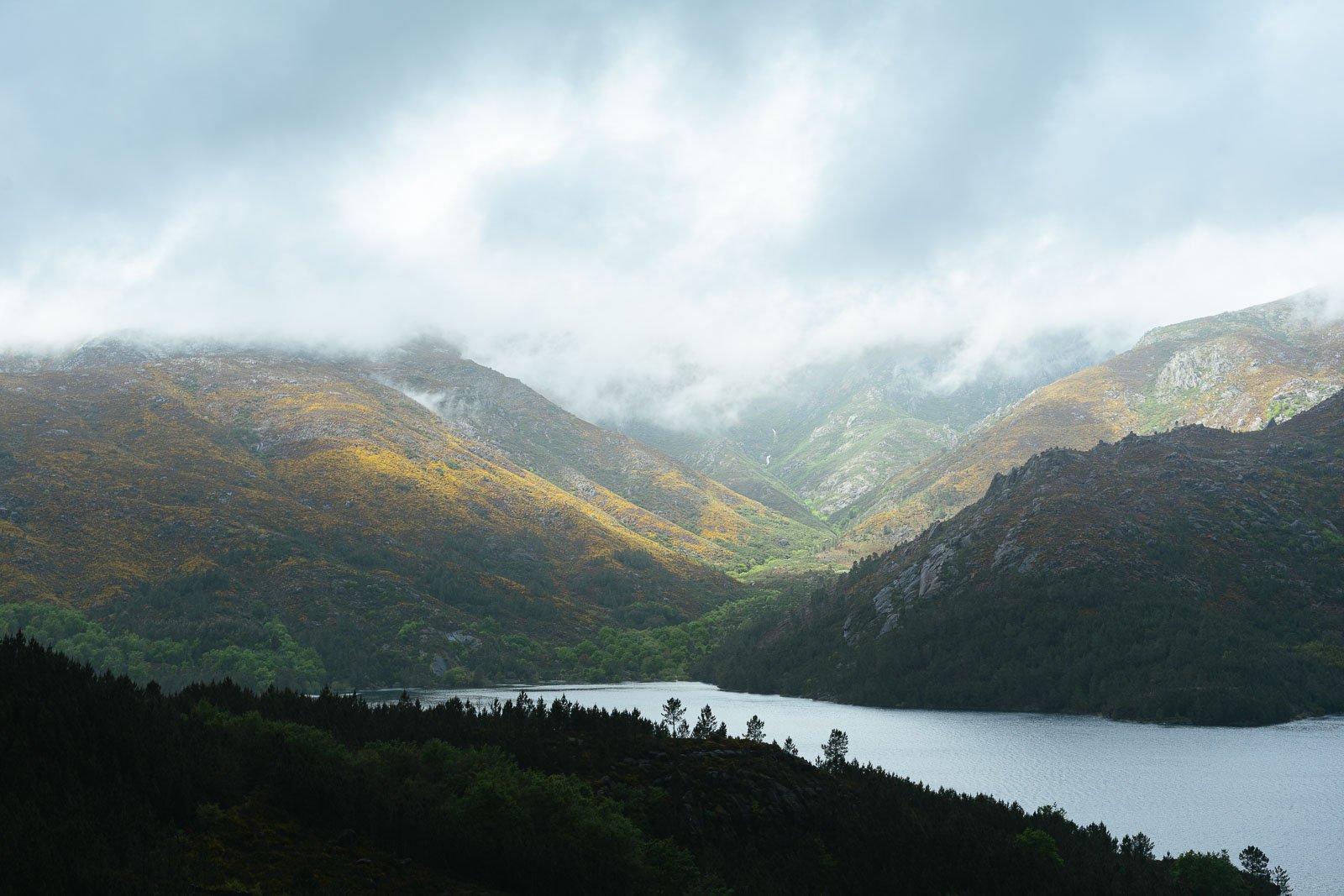 Montanhas verdes com um lago ao fundo, céu nublado com nuvens cobrindo o topo das montanhas.