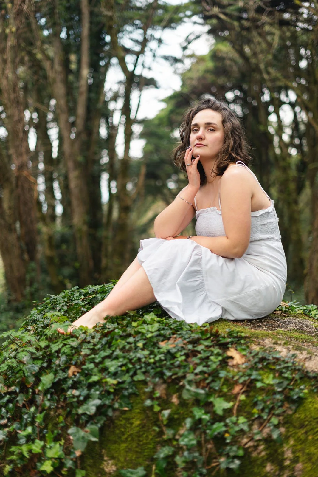 Mulher com cabelo castanho cacheado sentada em tronco de árvore coberto de hera em floresta, vestindo vestido branco, olhando para a câmera.