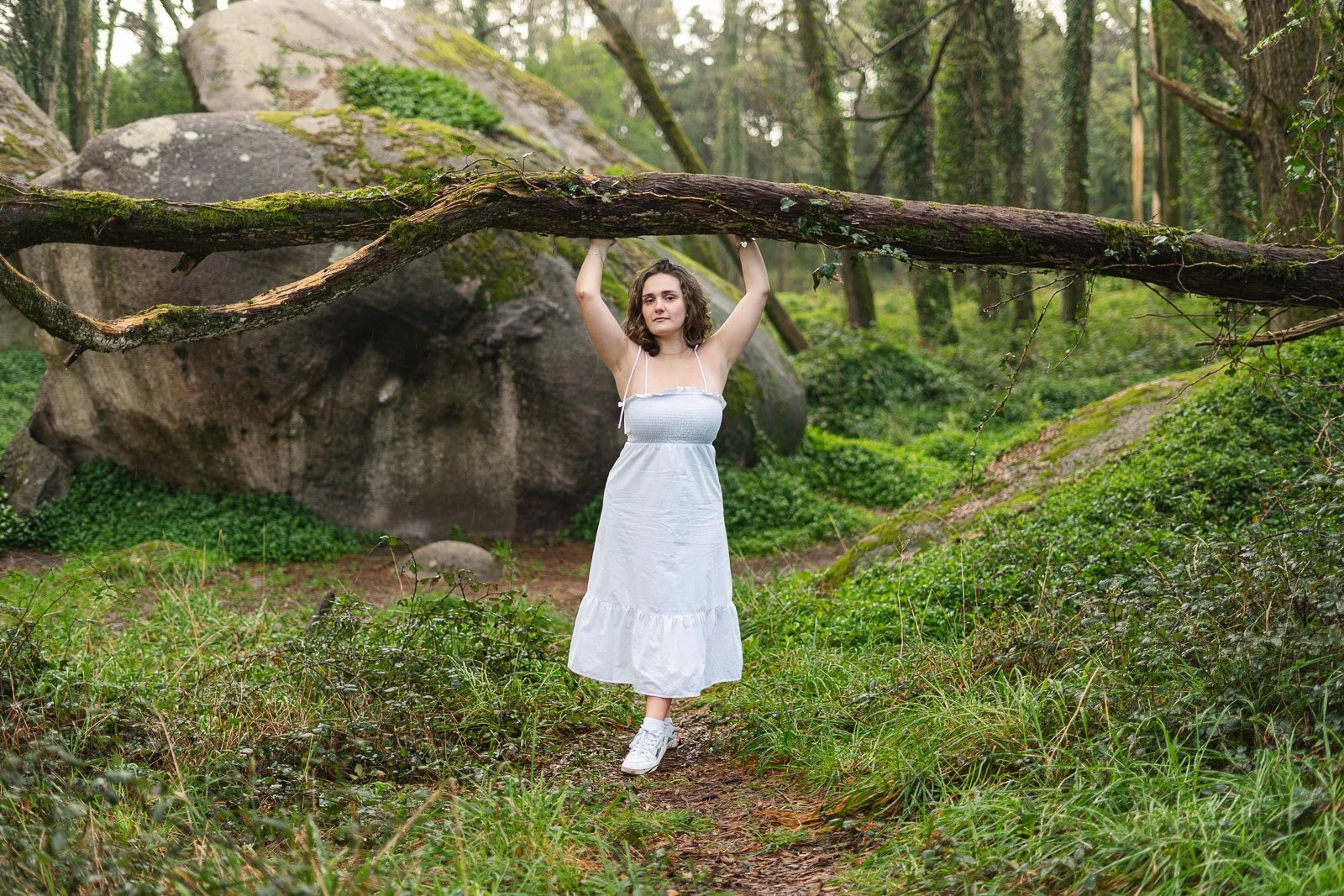Mulher jovem com cabelo cacheado, vestida com vestido branco, segurando um galho de árvore em um ambiente de floresta, ao lado de uma grande pedra.