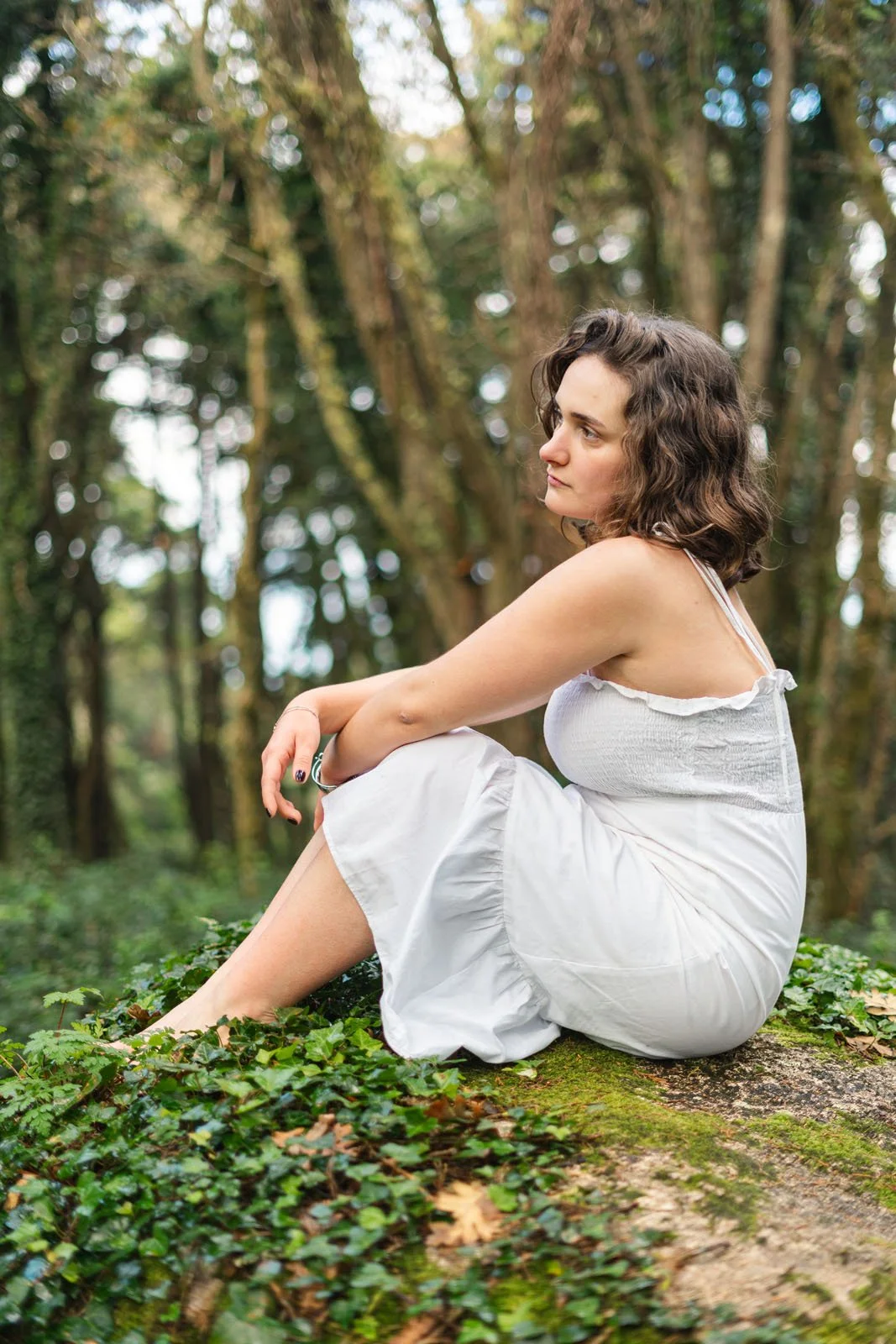 Mulher de cabelo castanho ondulado, vestida com vestido branco, sentada sobre um tronco de árvore coberto de musgo, em uma floresta, olhando para o lado.