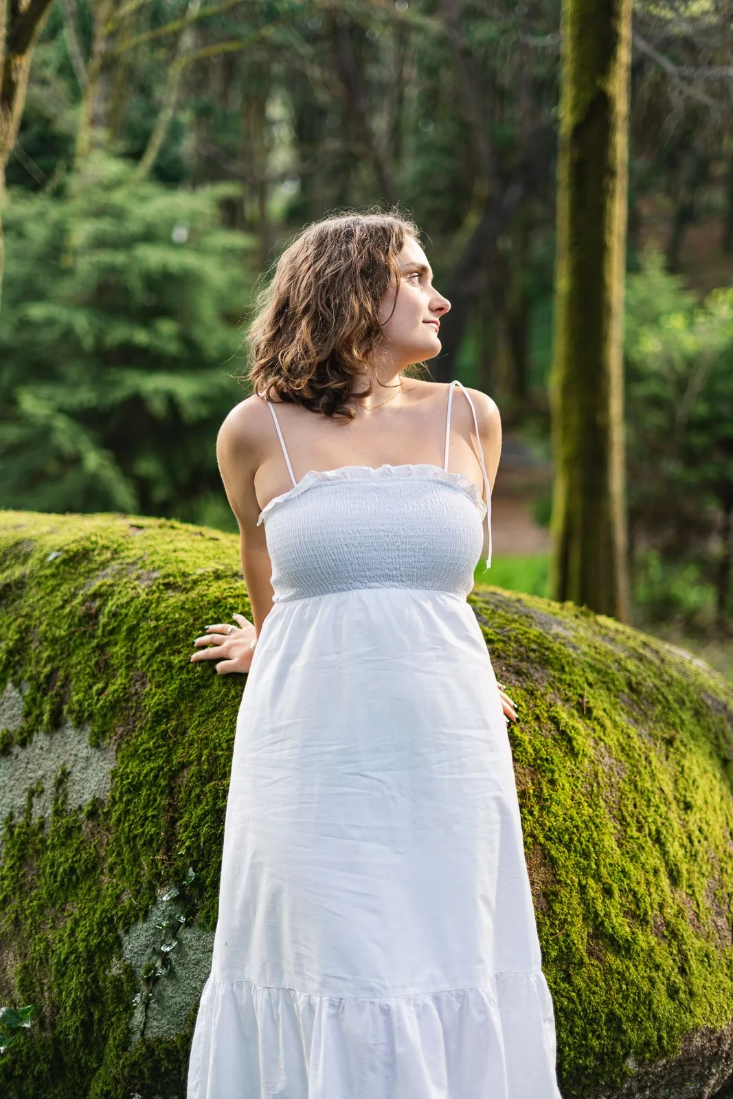 Jovem mulher com vestido branco posando ao ar livre em meio à floresta, apoiada em uma pedra coberta de musgo.