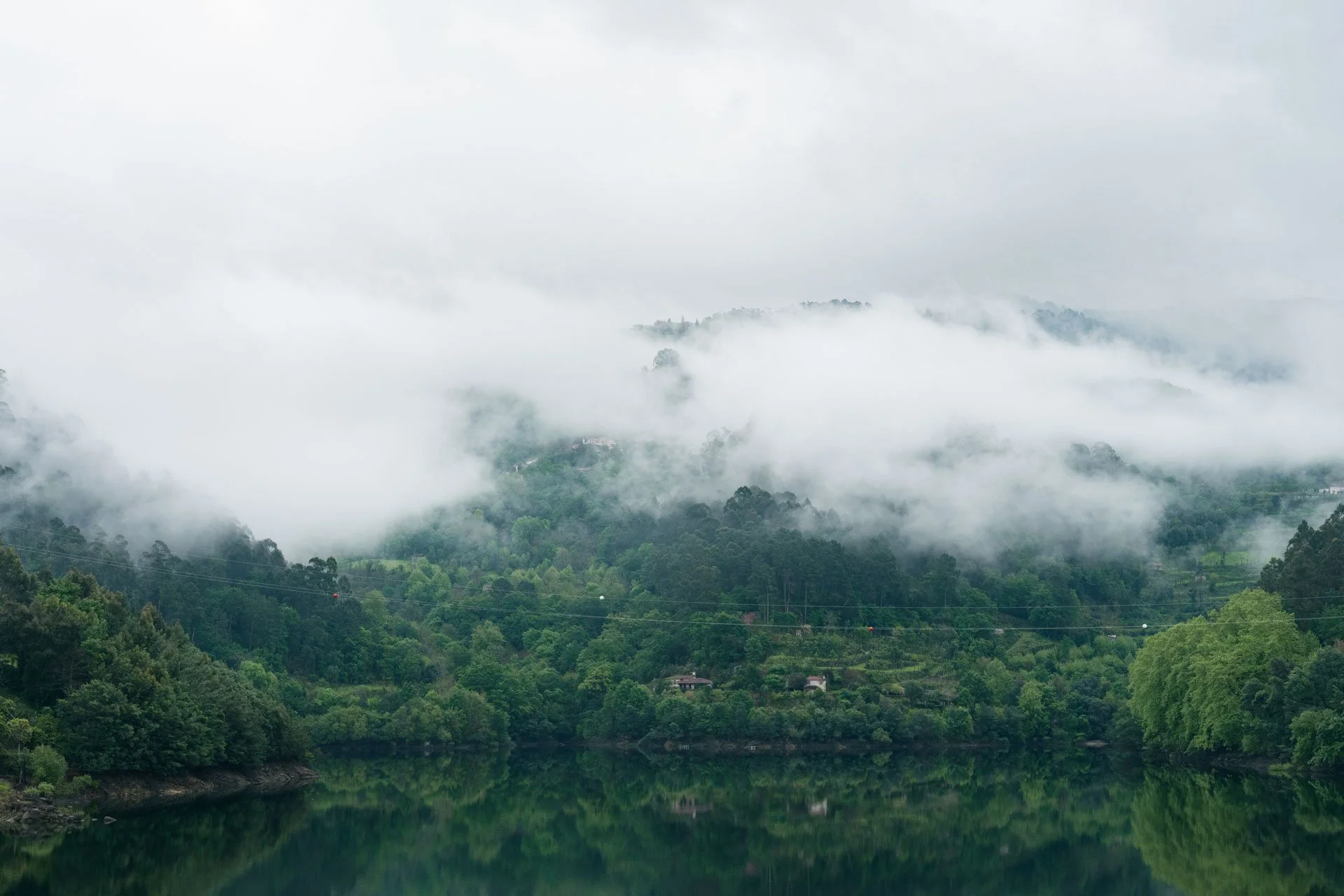 Paisagem de uma floresta exuberante ao redor de um lago tranquilo, com montanhas ao fundo cobertas por nuvens e neblina.