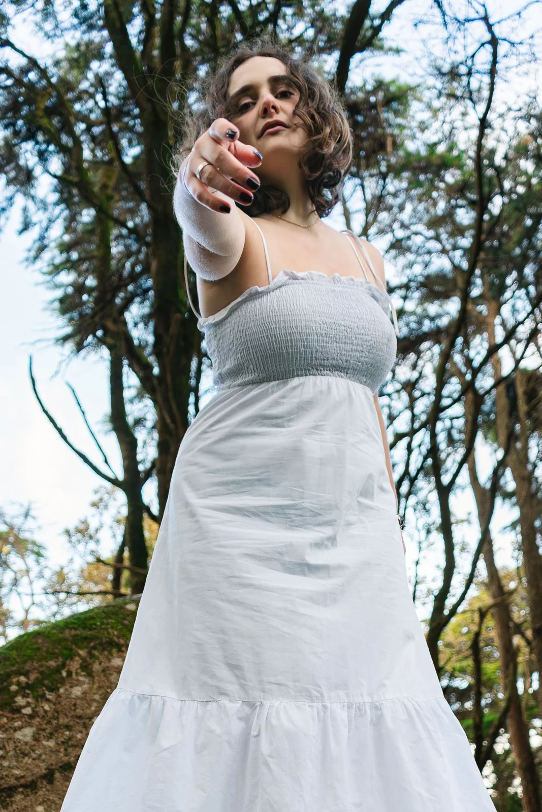 Mulher com cabelo cacheado, vestindo vestido branco, apontando para a câmera em uma floresta com árvores ao fundo.