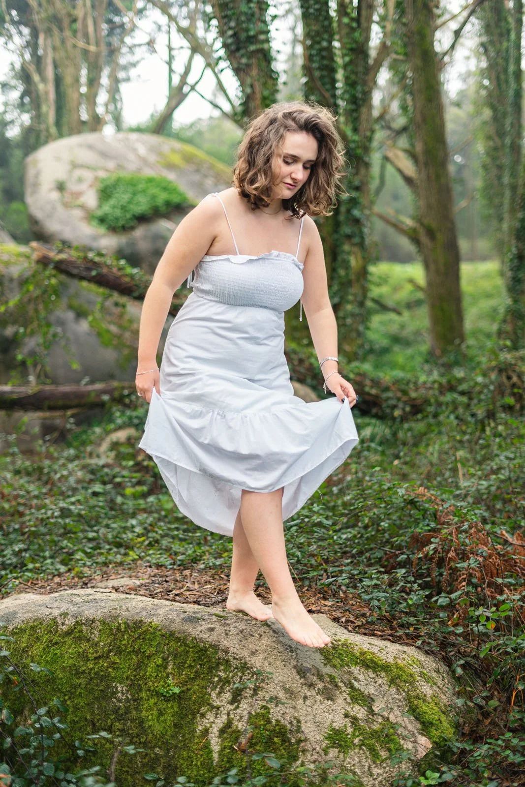 Mulher com cabelo cacheado, vestindo vestido branco, caminhando descalça sobre uma pedra no meio de uma floresta.