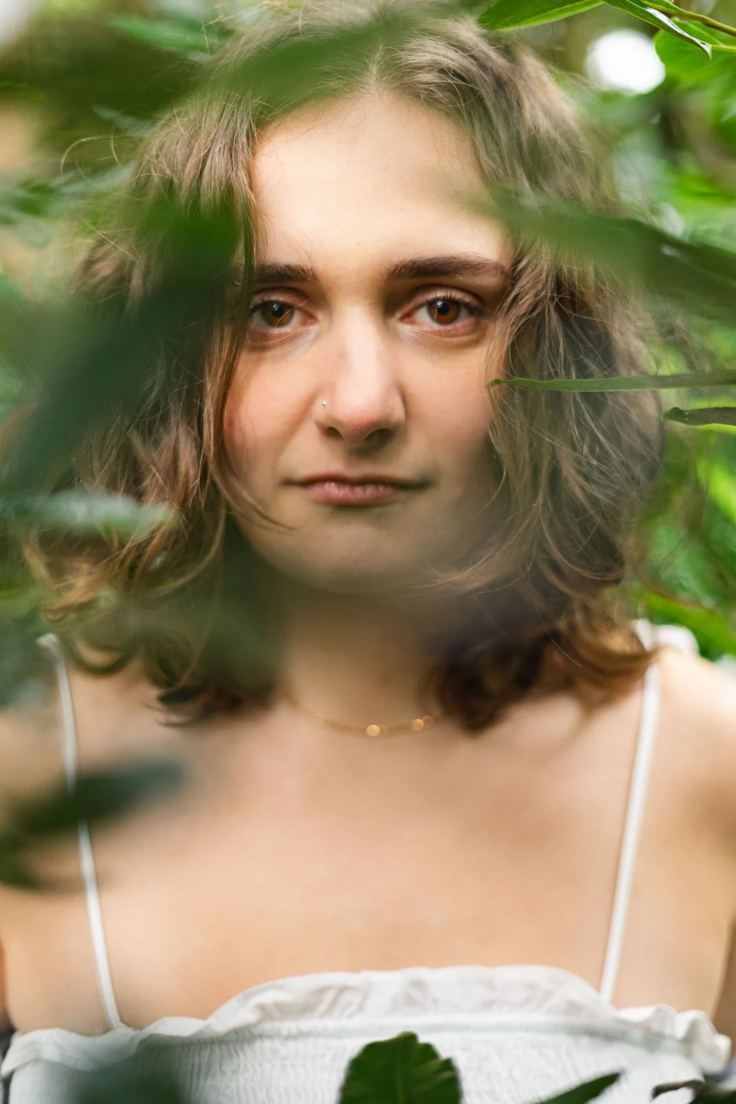 Mulher com cabelo castanho e olhos claros, usando vestido branco, cercada por folhas verdes.