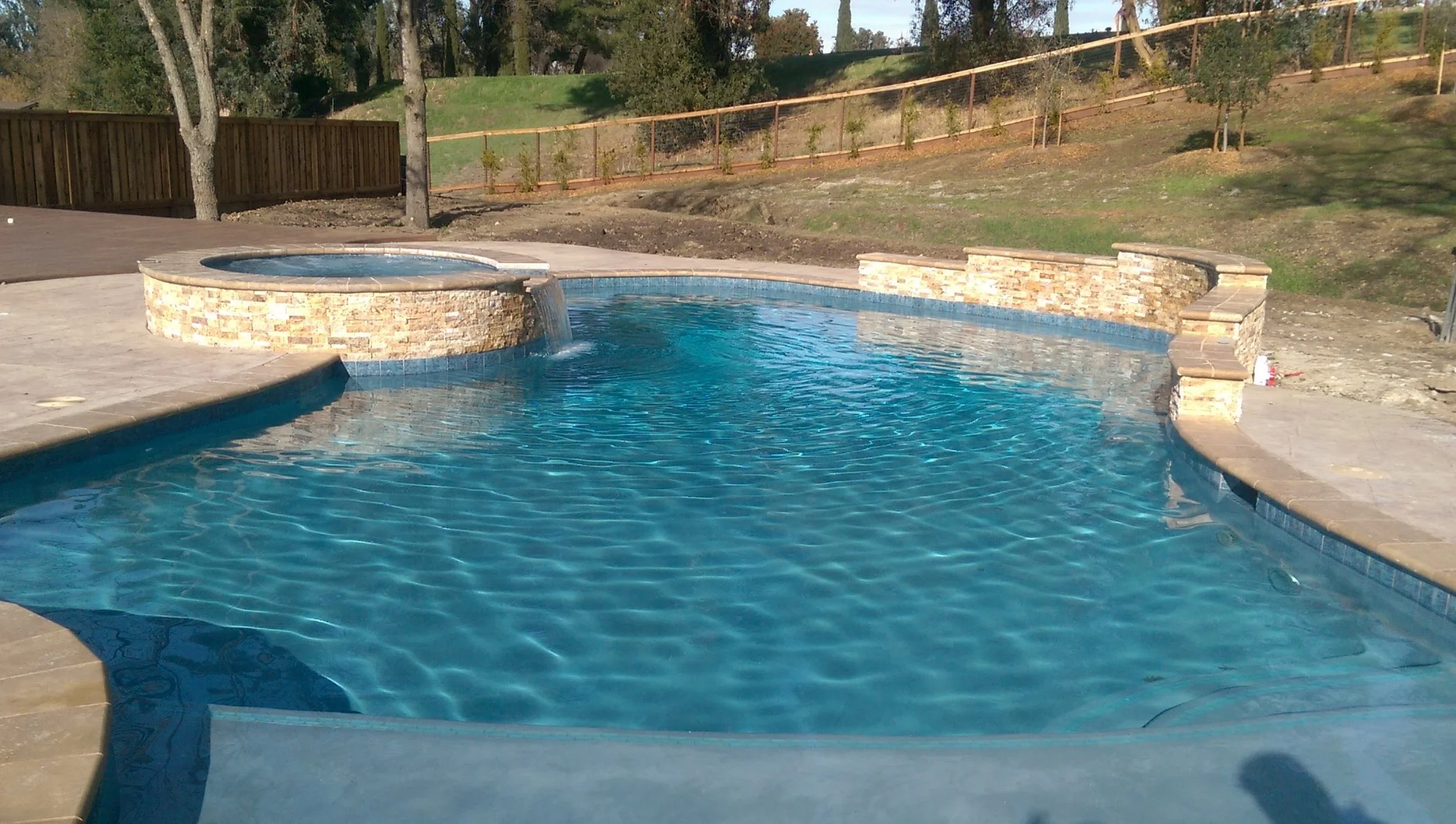 A backyard swimming pool with a hot tub, surrounded by a paved deck and a wooden fence, with trees and a hill in the background.