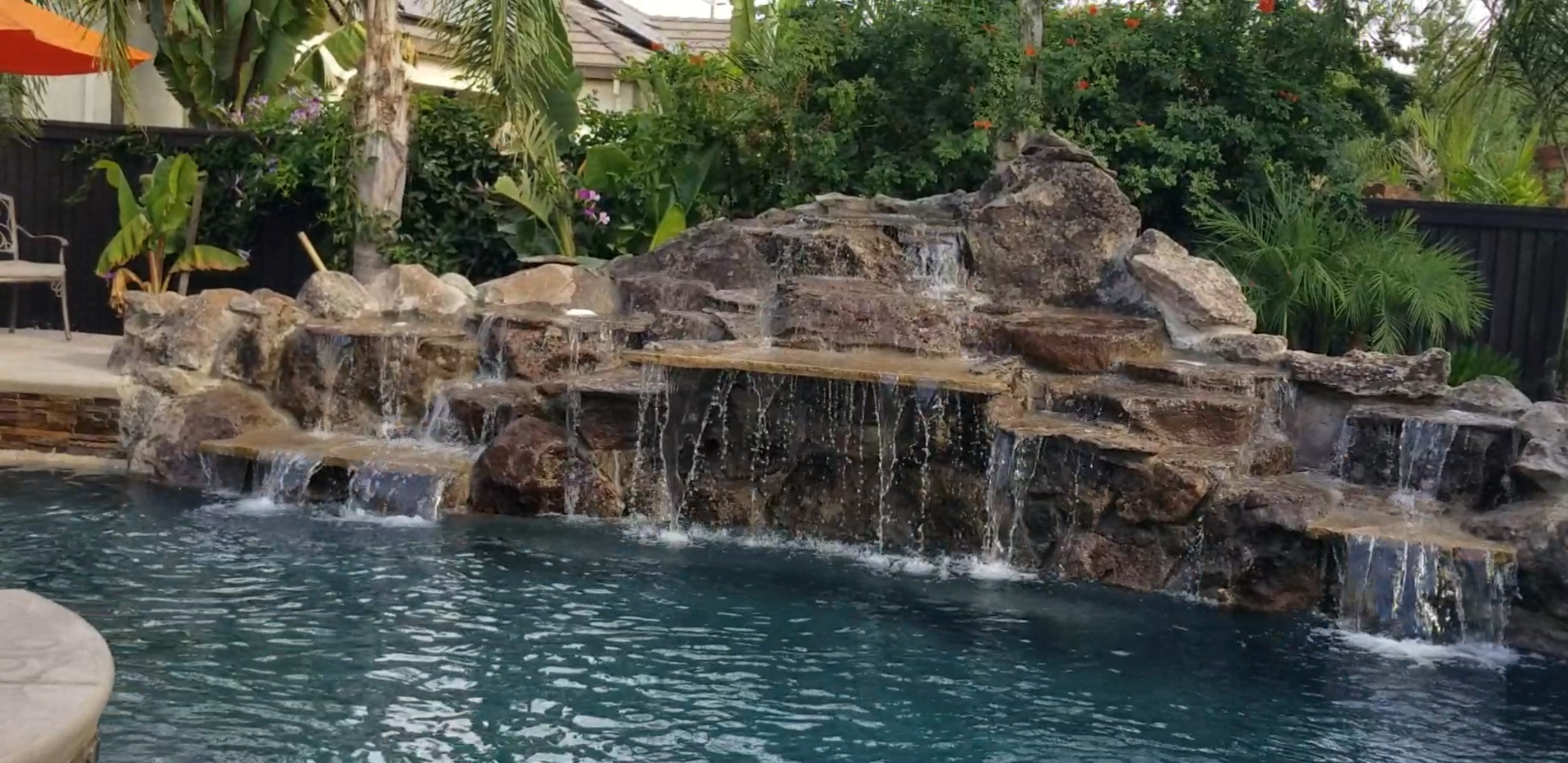 A backyard with a water feature and waterfall flowing over rocks into a swimming pool, surrounded by lush green plants and trees.