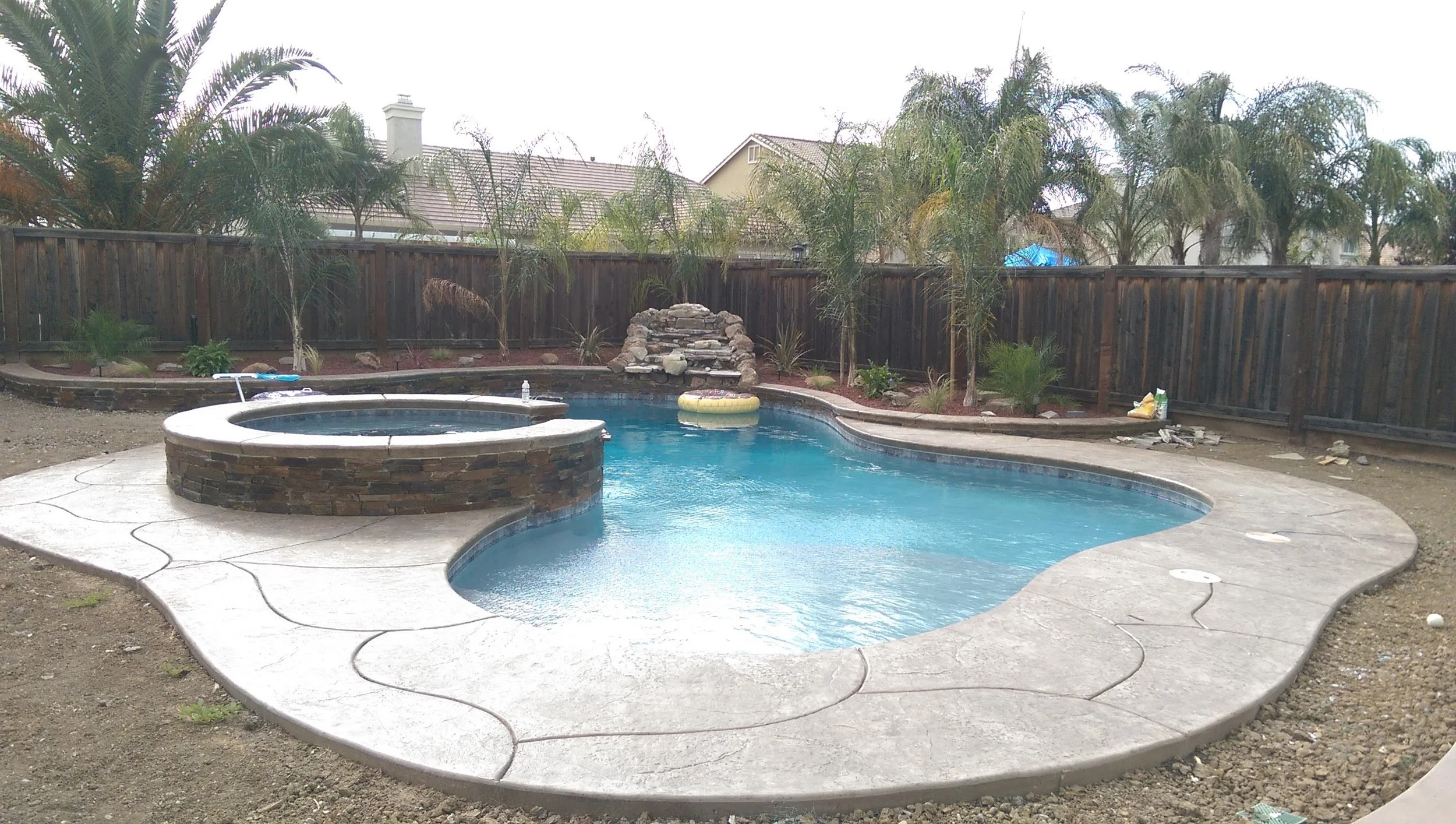 Backyard swimming pool with a hot tub, fountain, and a rock waterfall feature, surrounded by a wooden fence and landscaping with trees and plants.