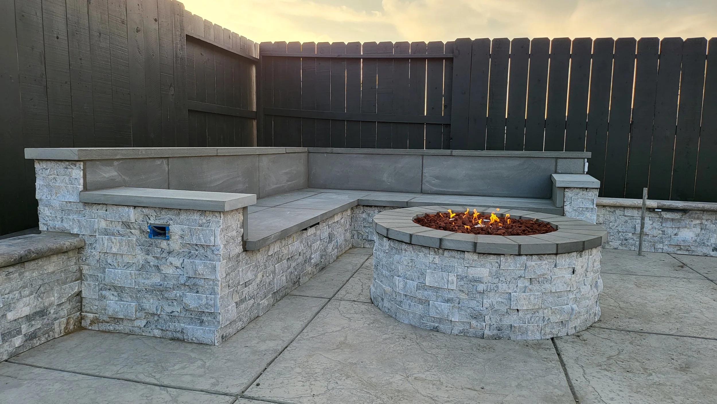 Backyard patio with built-in stone seating, a fire pit with flames, and a tall wooden privacy fence, during sunset.