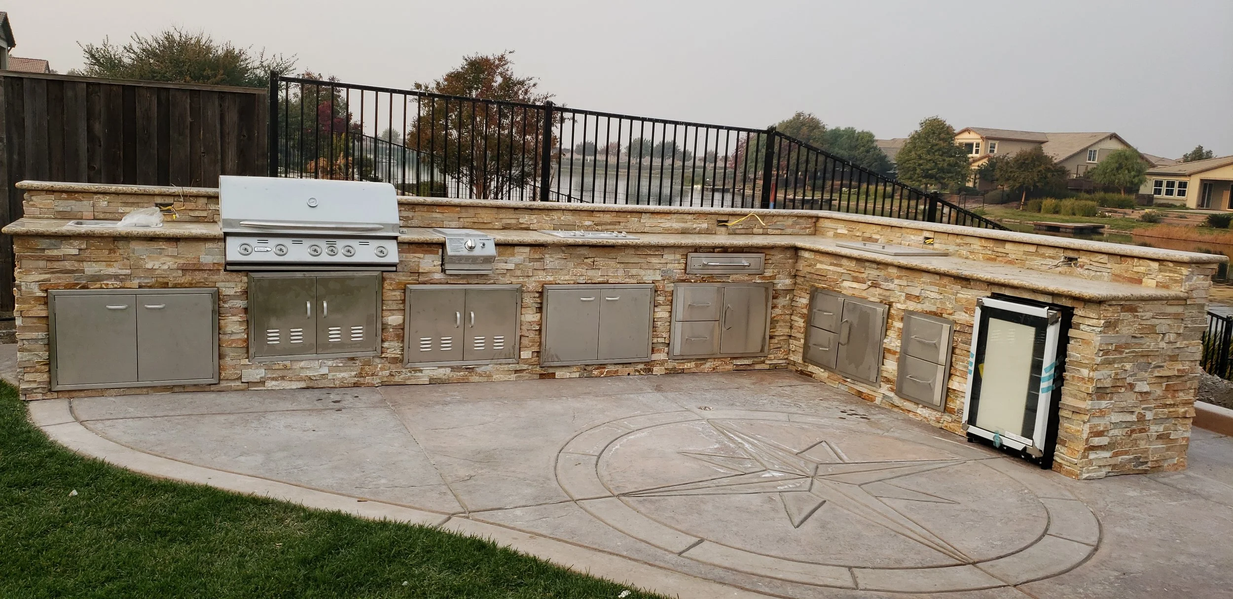 An outdoor kitchen with a stone counter and stainless steel cabinets, a built-in grill, and a small refrigerator, set on a concrete patio with an emblem design in the concrete.
