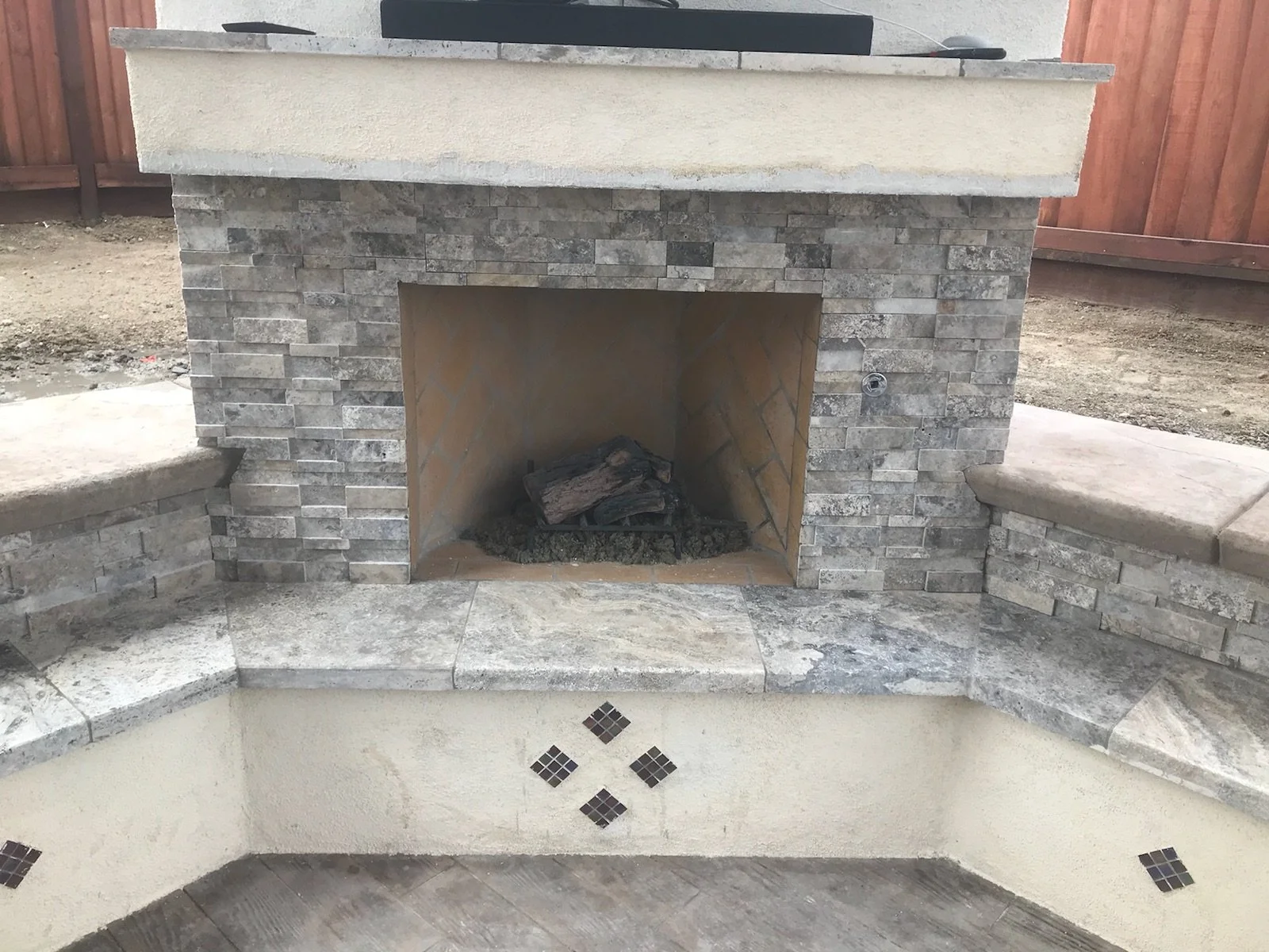 Outdoor stone fireplace with a stone surround, built into a concrete and stucco base, with a few logs inside the fireplace. A wooden privacy fence and dirt ground are visible in the background.