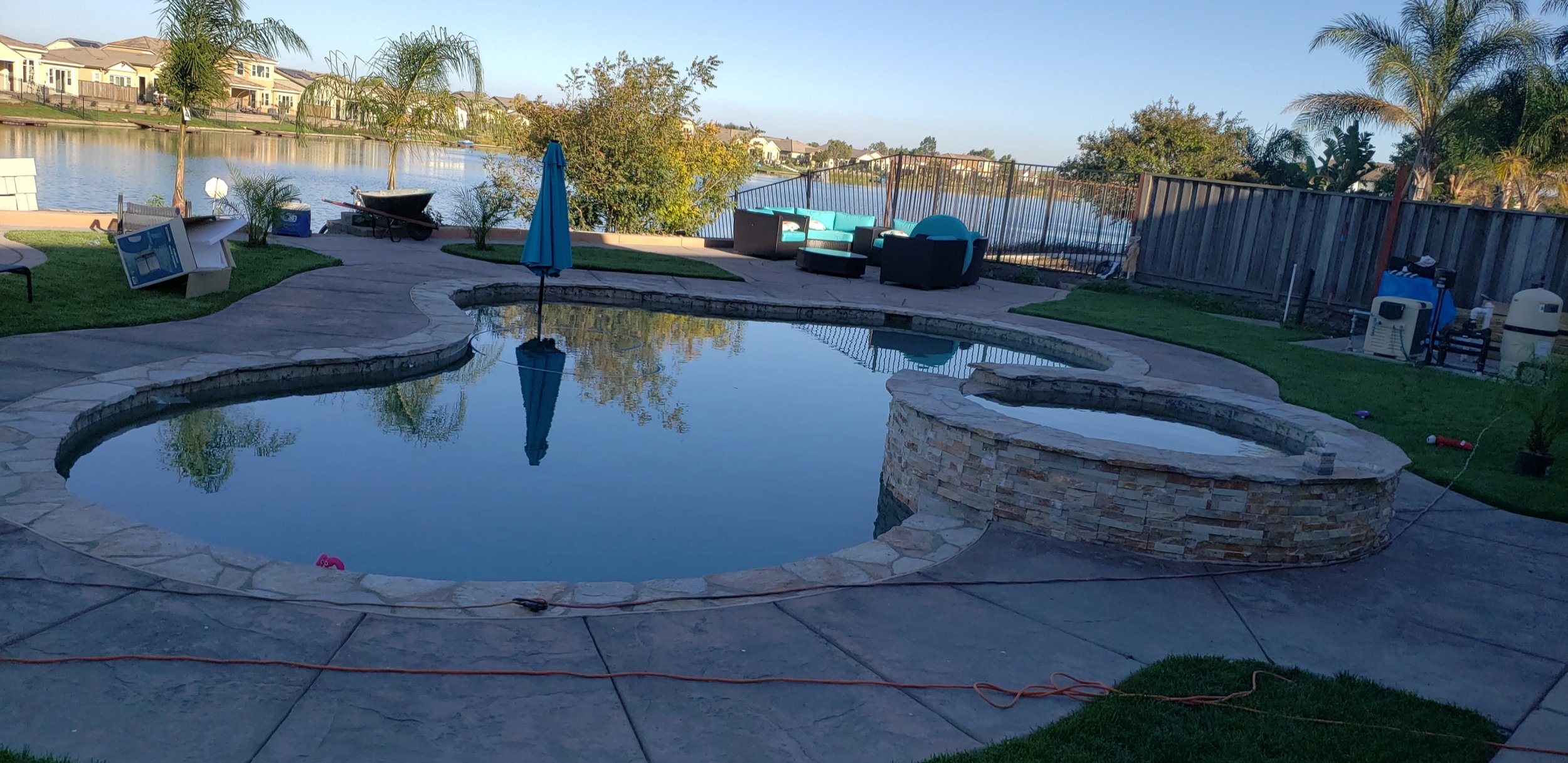 Backyard with a kidney-shaped swimming pool, a hot tub with a stone exterior, poolside furniture including a turquoise and black cushioned seating area, a blue umbrella, and a waterfront view with houses across the lake. There are some tools and equi
