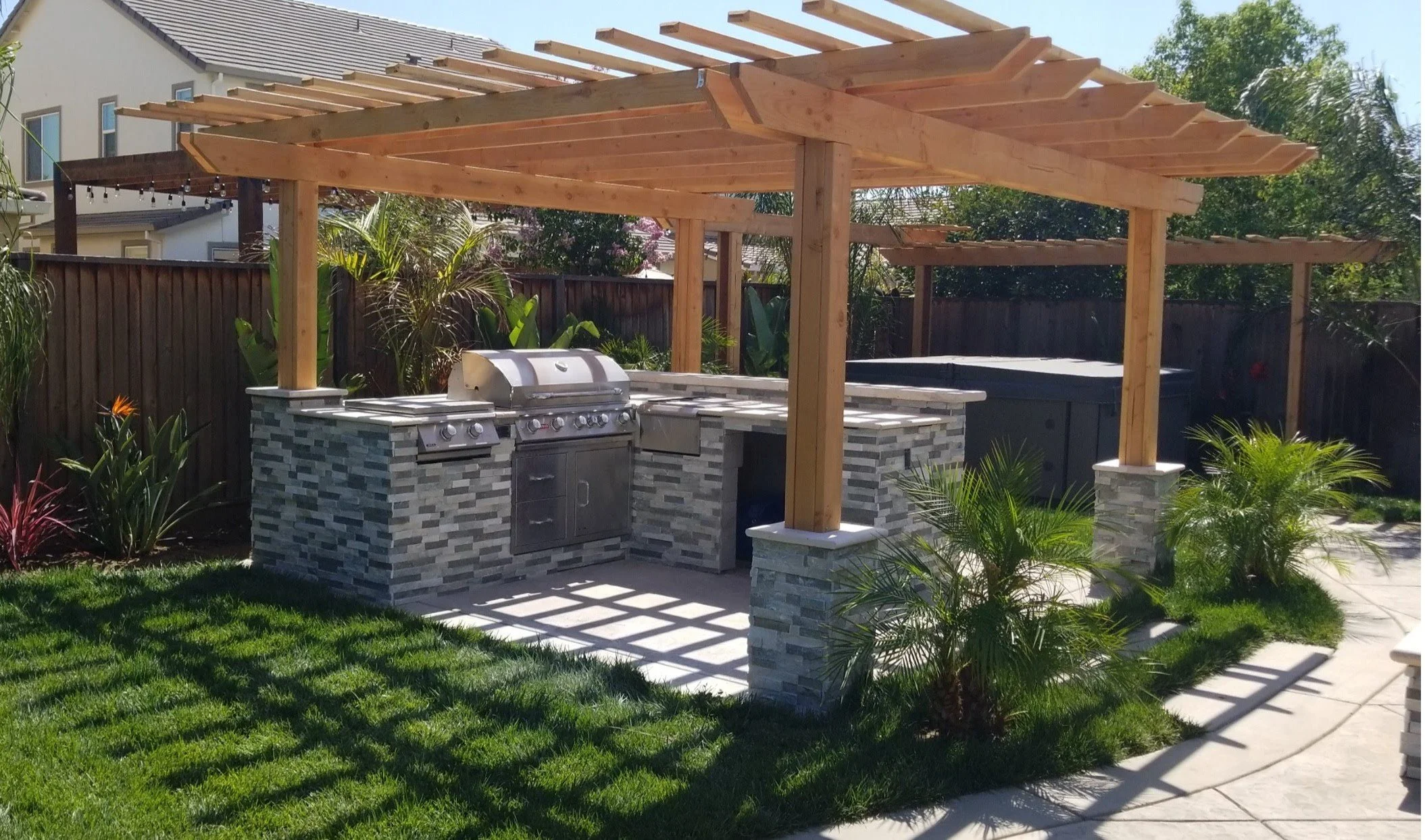 Outdoor backyard kitchen with stone countertop, built-in grill, and wooden pergola, surrounded by green plants and a grassy lawn.