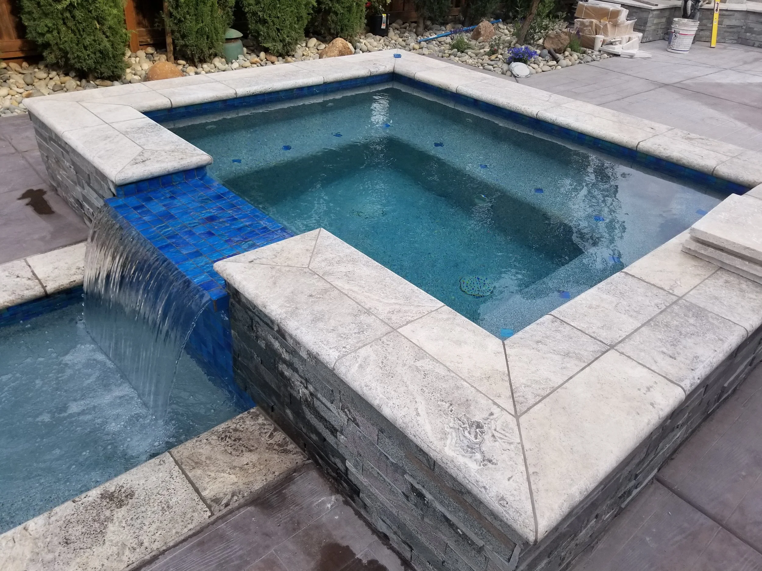 A square hot tub with a stone and brick exterior, featuring a small waterfall with blue mosaic tiles, situated outdoors on a patio with construction supplies visible in the background.