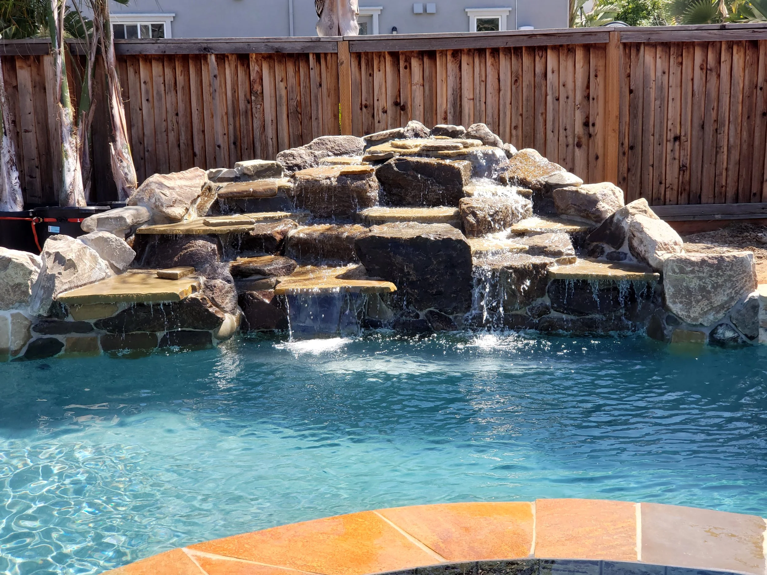 Backyard swimming pool with a rock waterfall feature, surrounded by a wooden fence.