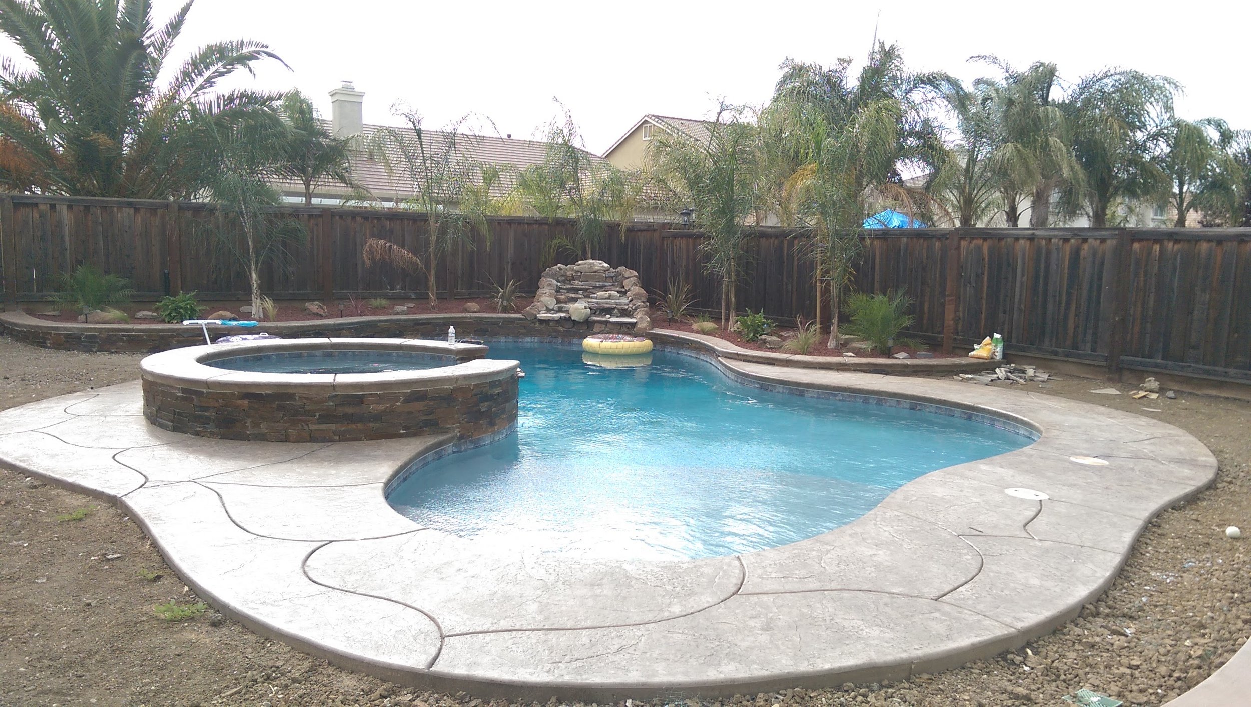 Backyard with a swimming pool and hot tub, surrounded by a wooden fence and trees.