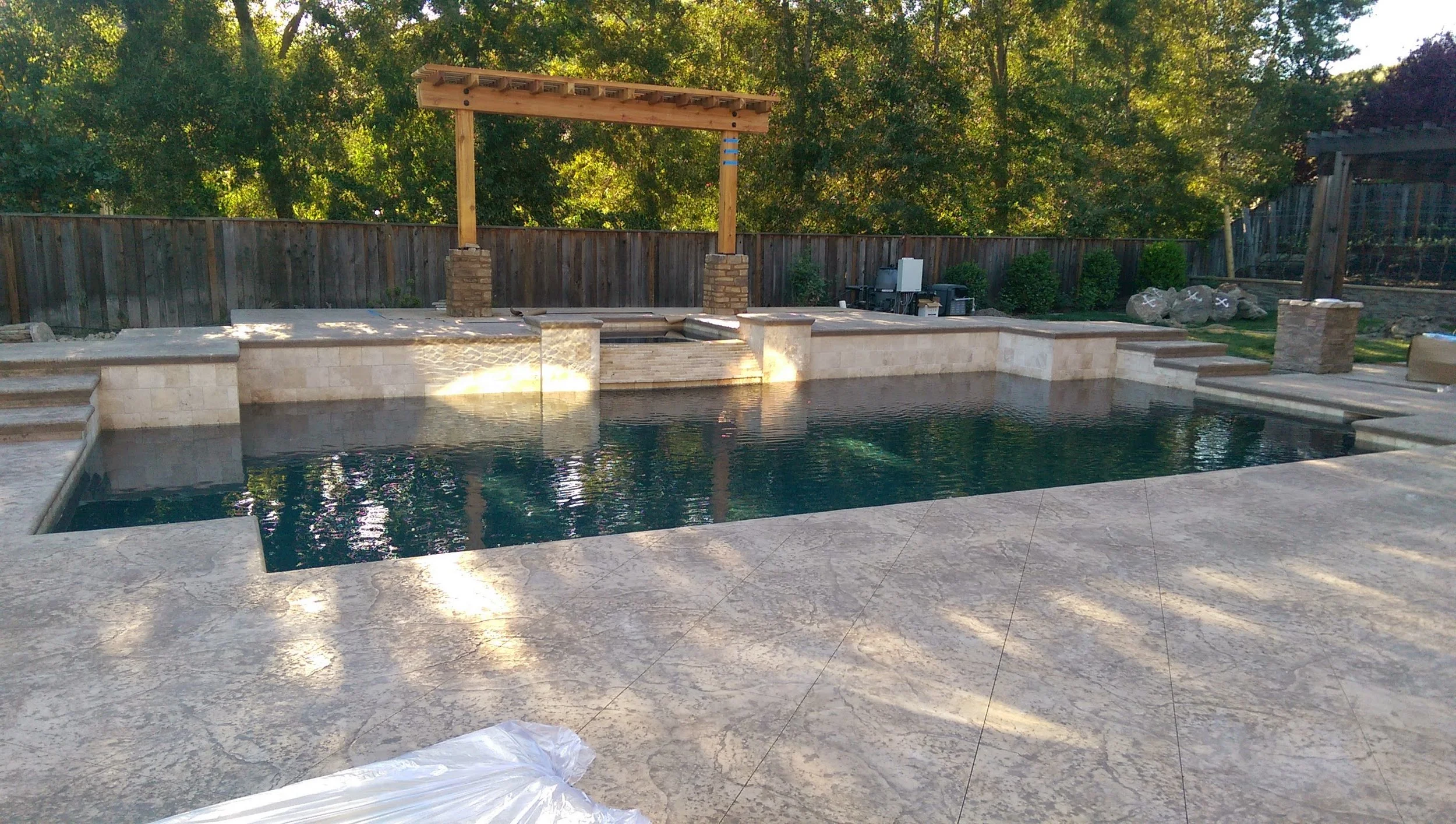 Finished backyard swimming pool with a stone deck, surrounded by a wooden privacy fence and landscaped with trees and bushes.
