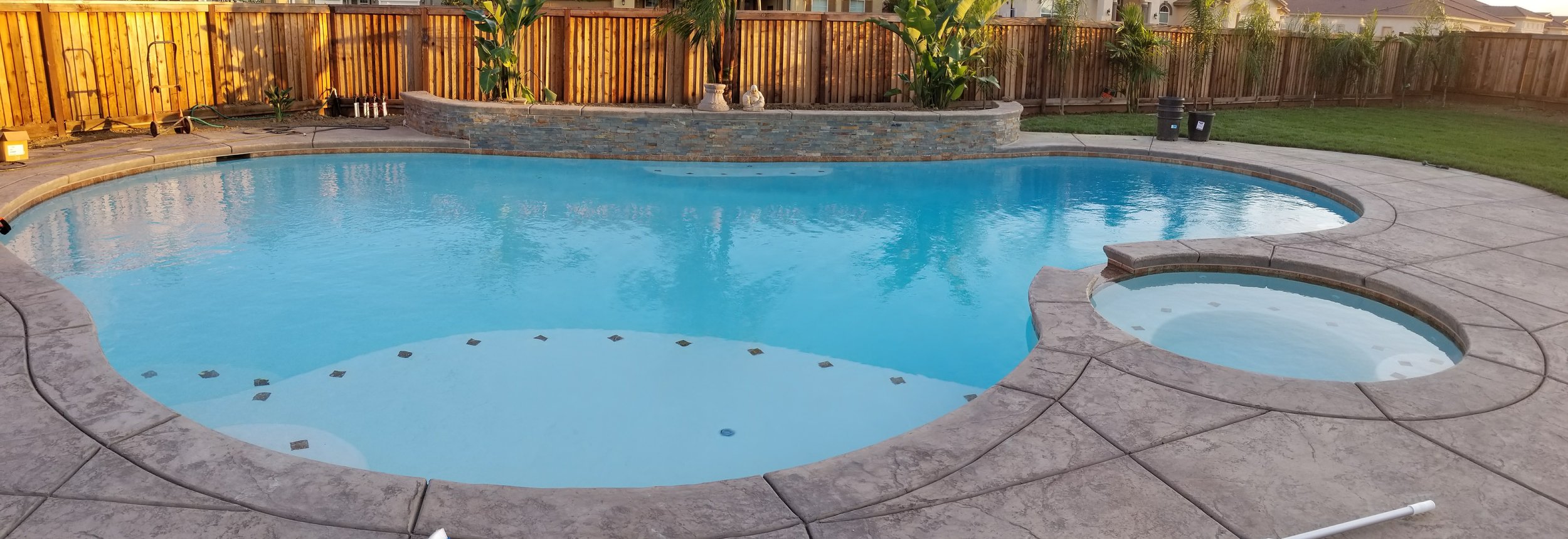 Backyard swimming pool with a small attached spa, surrounded by a concrete deck, wooden fence, and some plants and decorations on a sunny day.