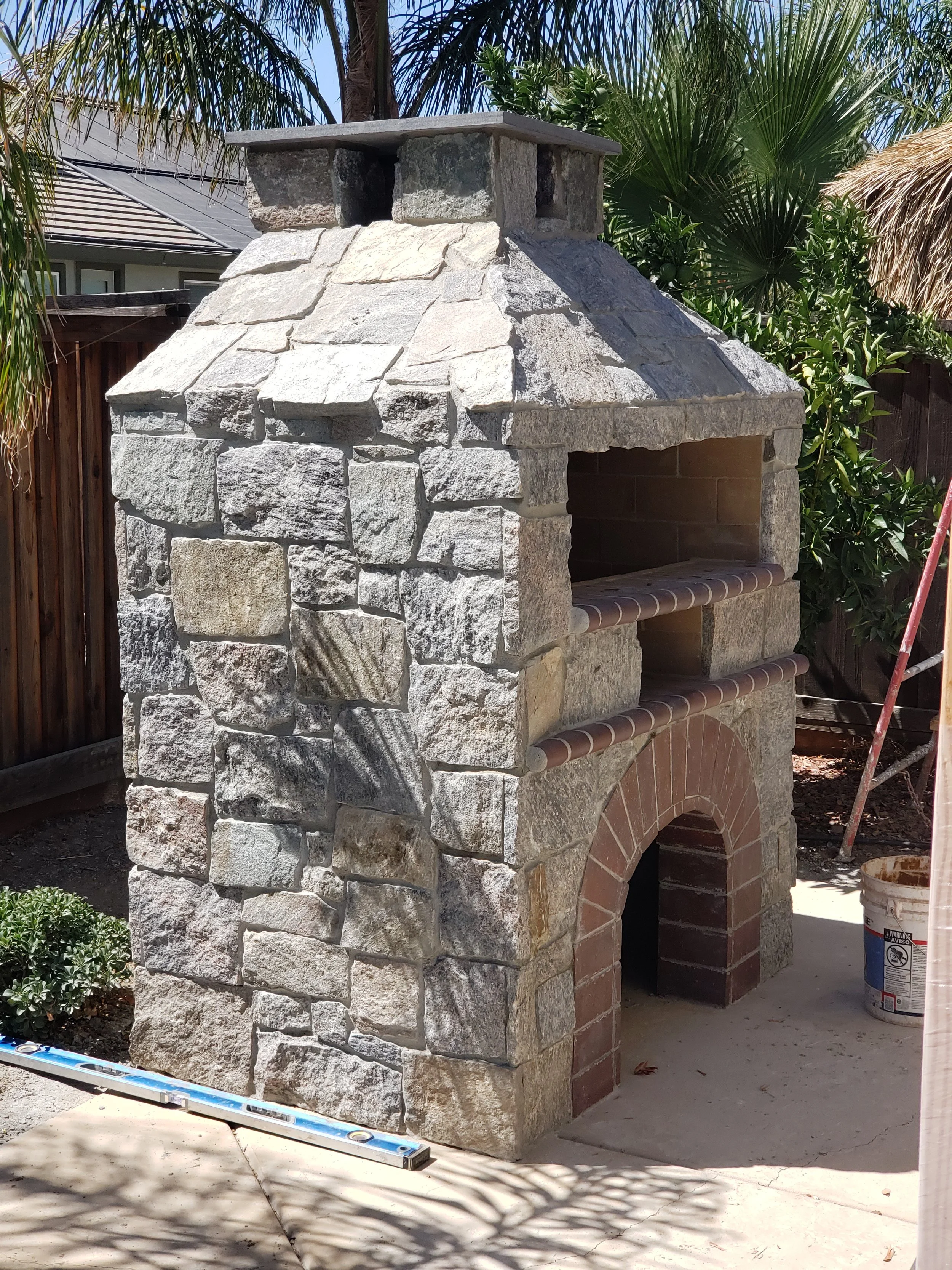 Stone outdoor fireplace with brick arch and tiled shelves, surrounded by desert plants and a wooden fence.