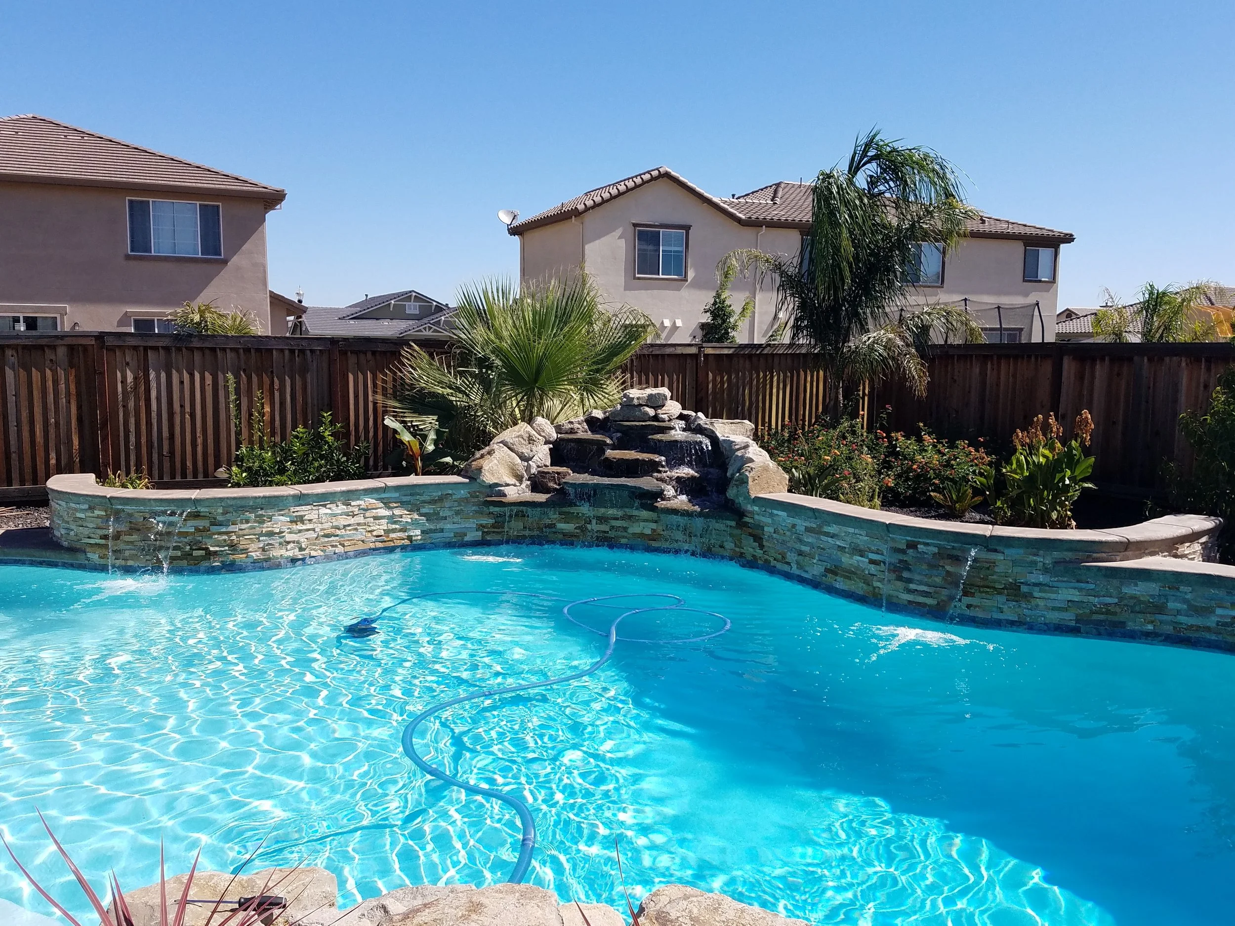 Backyard swimming pool with a rock waterfall feature, surrounded by a wooden fence and plants, with neighboring houses and a clear blue sky in the background.