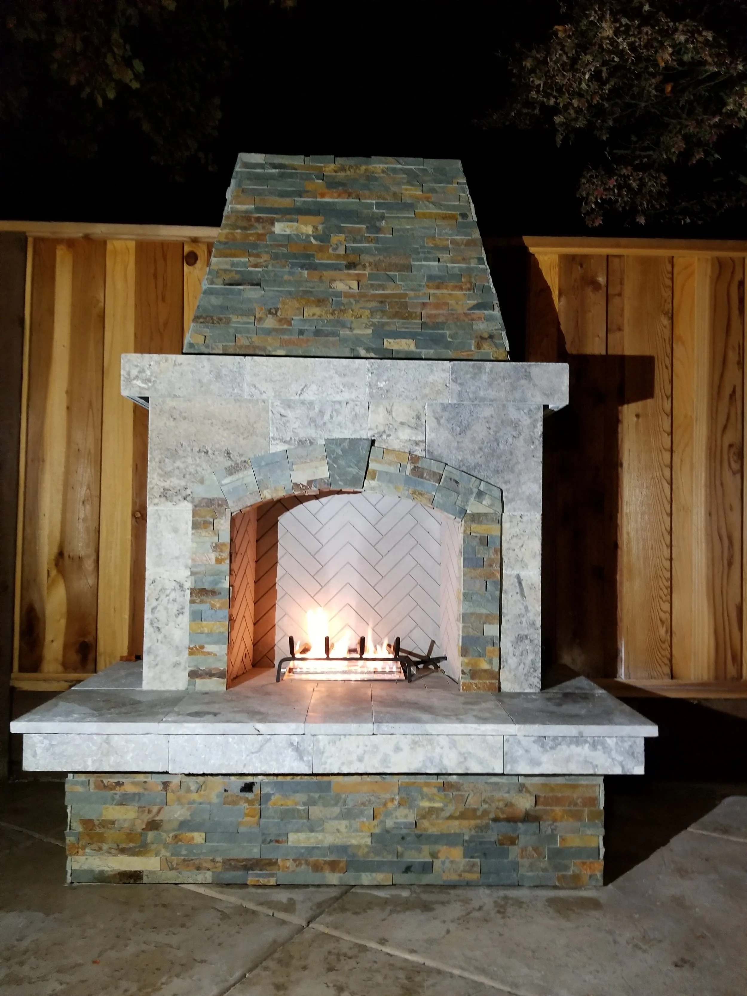 Outdoor stone fireplace with a fire burning, surrounded by a wooden fence at night.