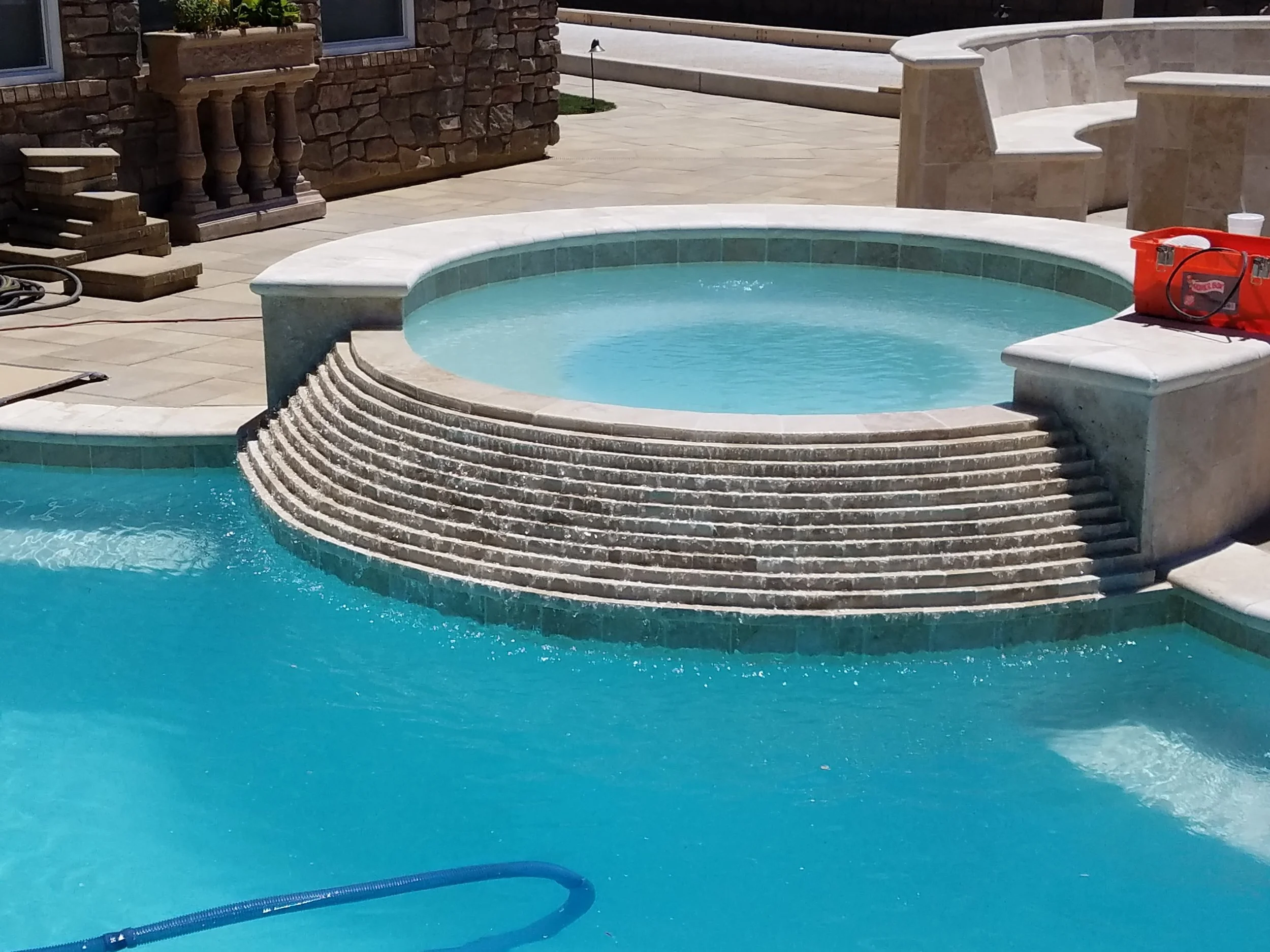 A swimming pool with a raised circular hot tub, steps leading up to it, and a surrounding patio area with outdoor furniture and a brick wall in the background.