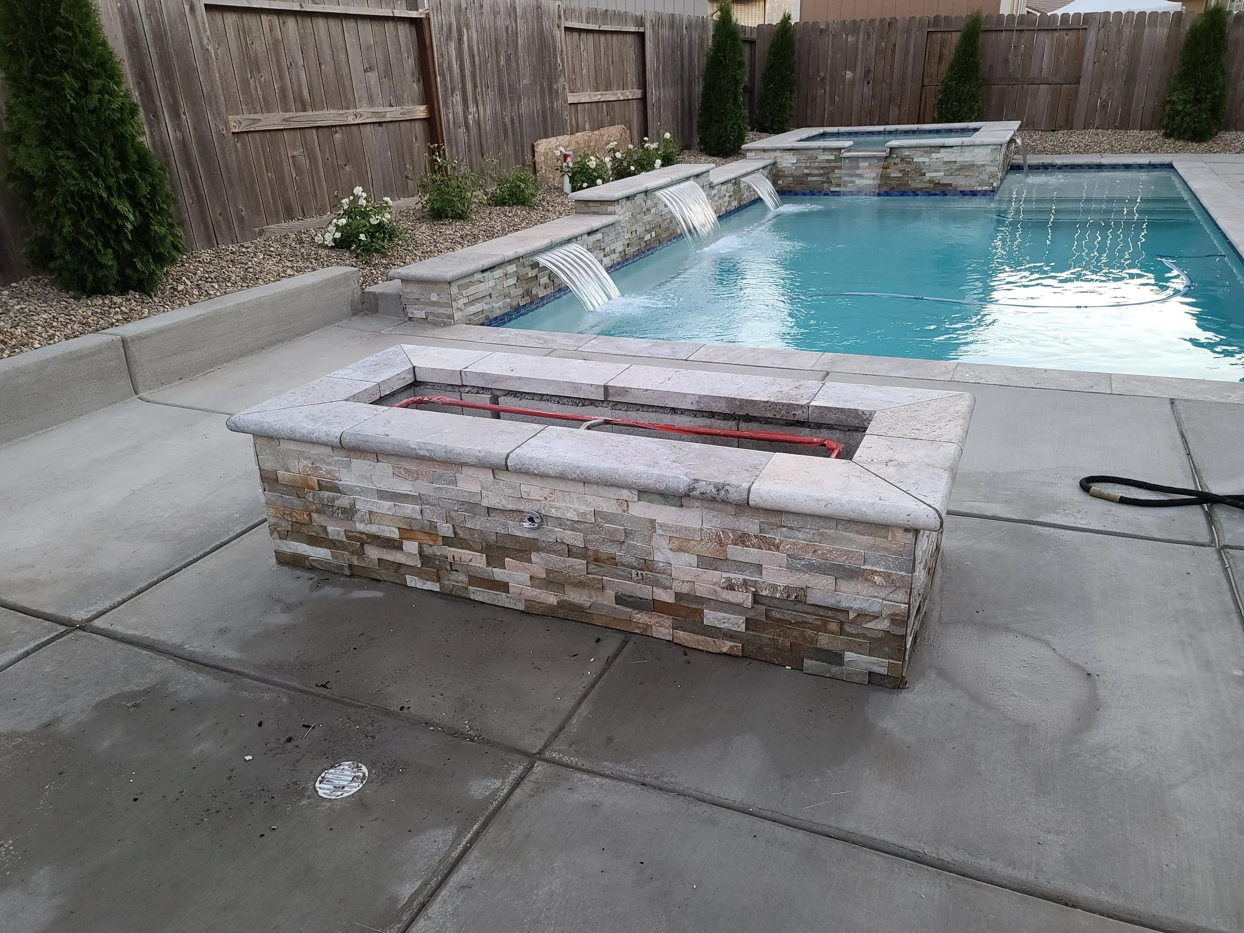 A backyard swimming pool with three water features, a small hot tub, and a wishing well in the foreground. The pool is surrounded by a concrete deck and a wooden fence with tall shrubs.