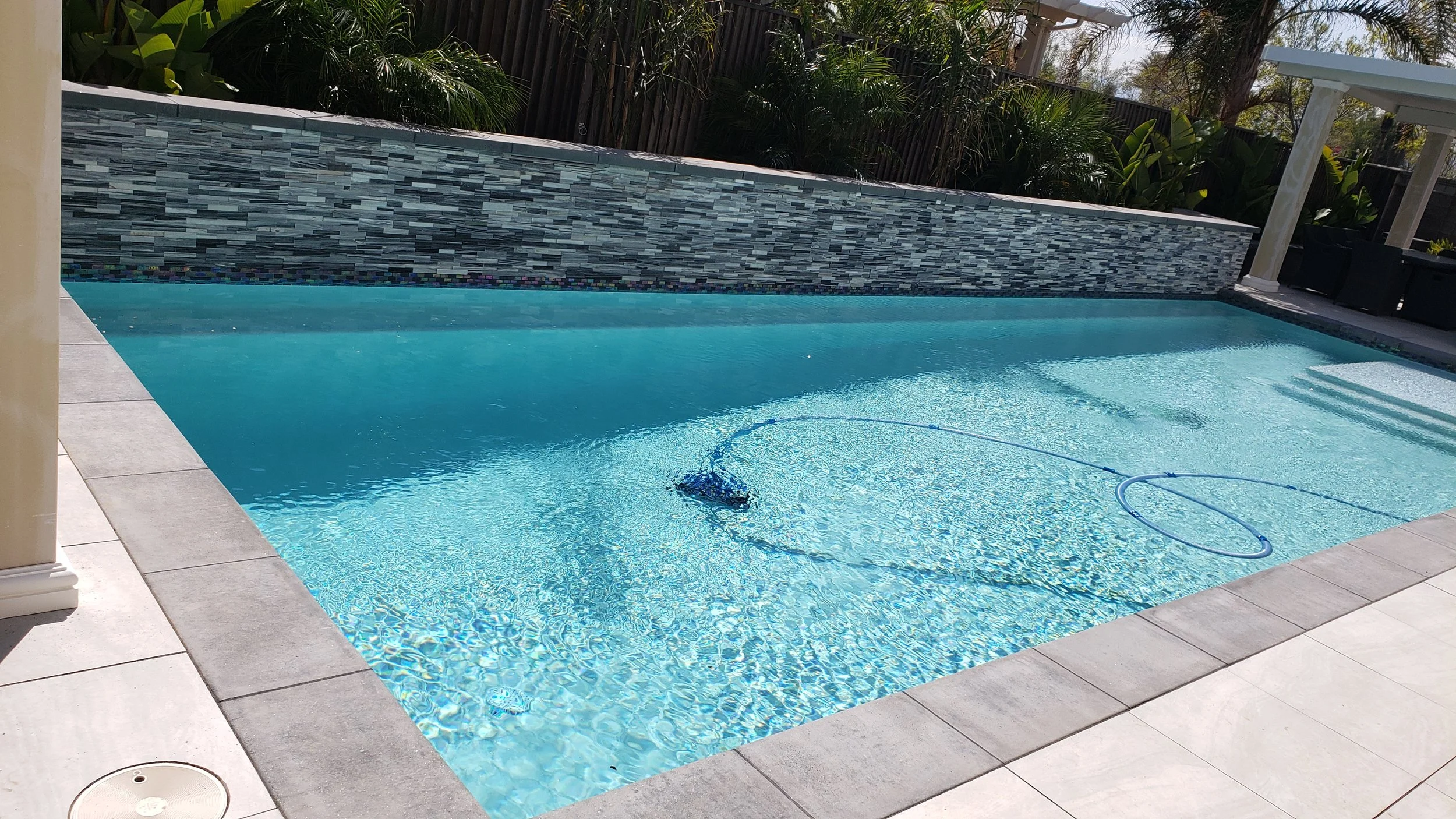 An in-ground swimming pool with clear blue water, a decorative tile wall at the far end, and a tiled deck surrounding the pool. There are plants and trees in the background, along with a pergola on the right side.