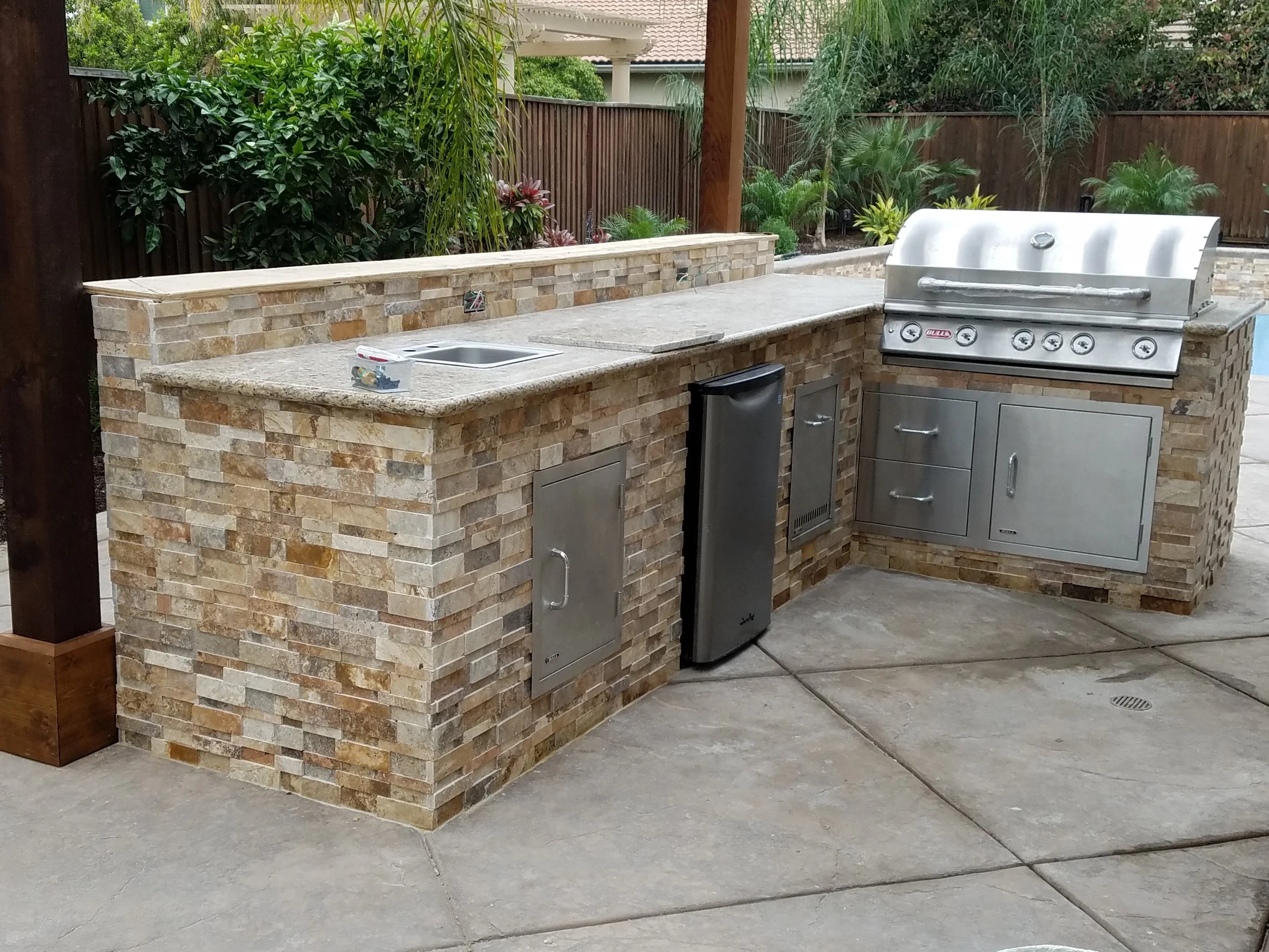 Outdoor kitchen with brick and stone counter, stainless steel grill, sink, small fridge, and storage cabinets, surrounded by a garden with plants and wooden fence.
