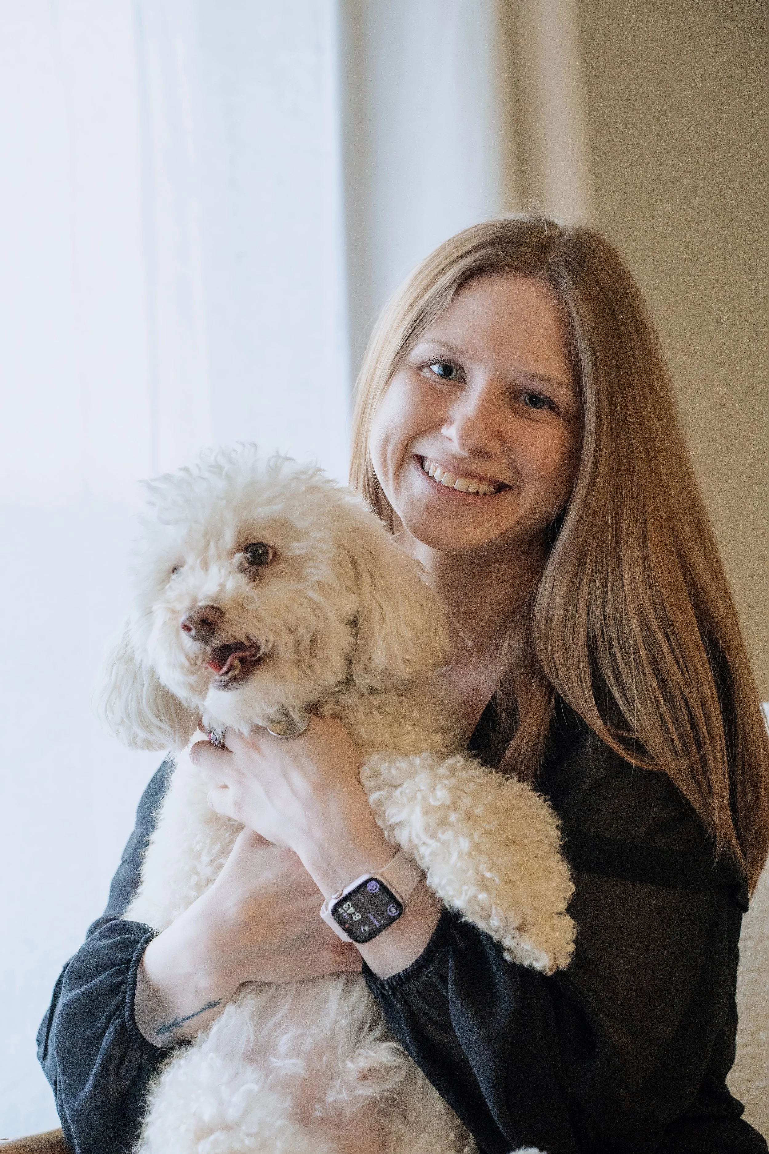 A woman with long red hair smiling and holding a fluffy cream-colored dog indoors near a window.