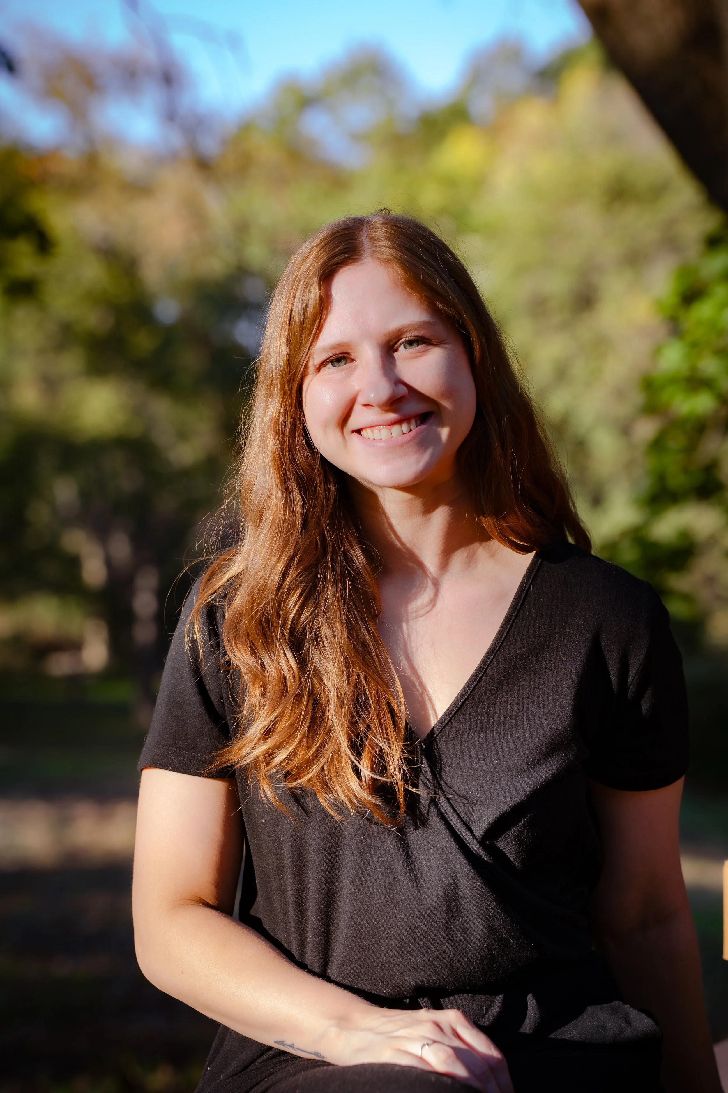 A young woman with long red hair smiling outdoors during daytime, wearing a black top with a v-neck, with trees and blue sky in the background.